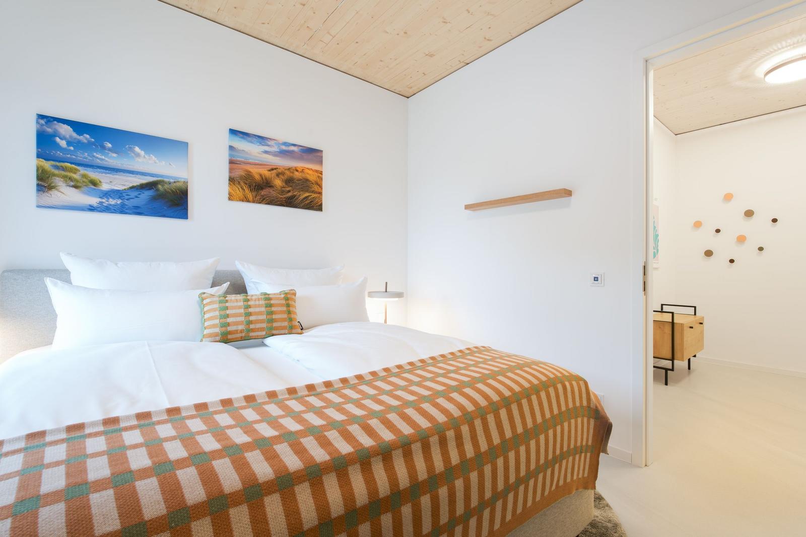 Schlafzimmer mit Bett, Wandbildern und Holzdecke. Tür führt in angrenzenden Raum.