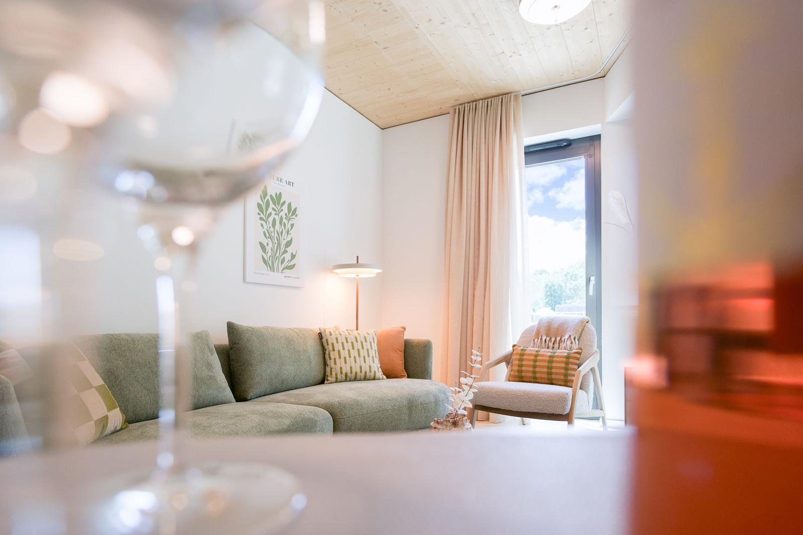 Wohnzimmer mit Sofa, Sessel und Fenster. Vordergrund: Weinflasche und Glas.