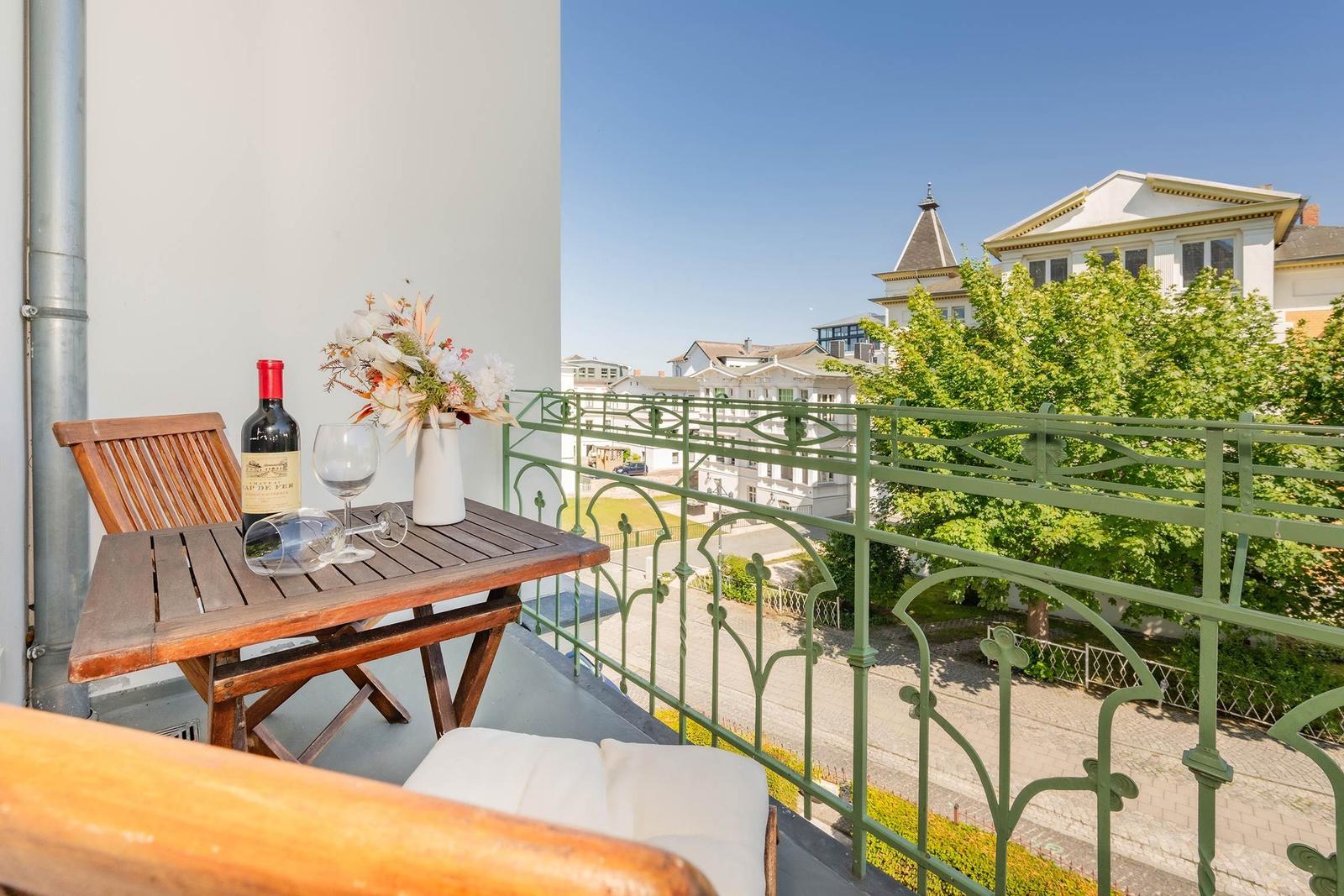 Balkon mit Tisch, Stuhl und Wein. Blick auf grüne Bäume und Gebäude.