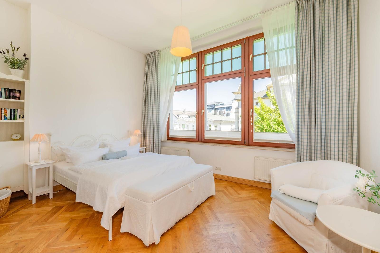 Schlafzimmer mit großem Fenster, weißem Bett und Sessel. Holzboden und Bücherregal.