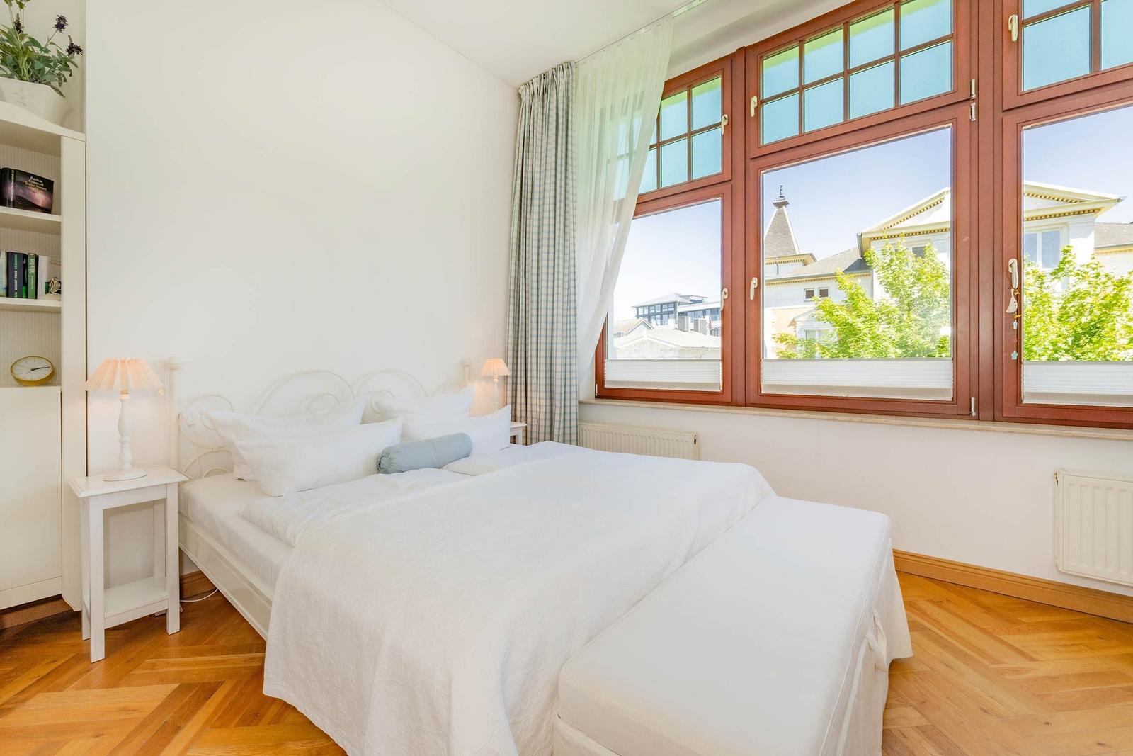 Schlafzimmer mit weißem Bett, Holzboden und großem Fenster mit Blick auf Gebäude.