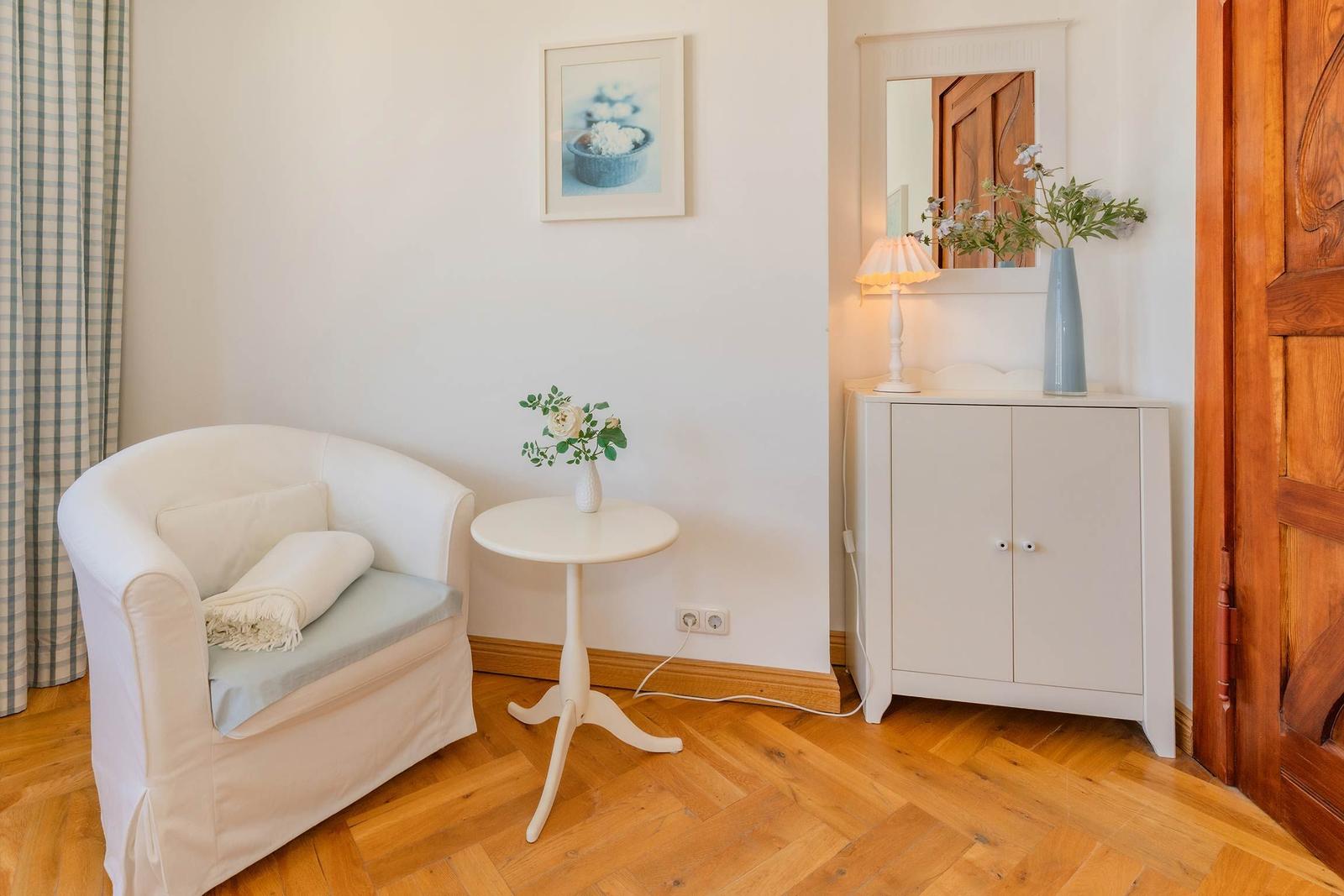 Ein weißer Sessel mit Kissen und Decke neben einem runden Tisch. Hinter ihm steht ein Schrank mit Spiegel.