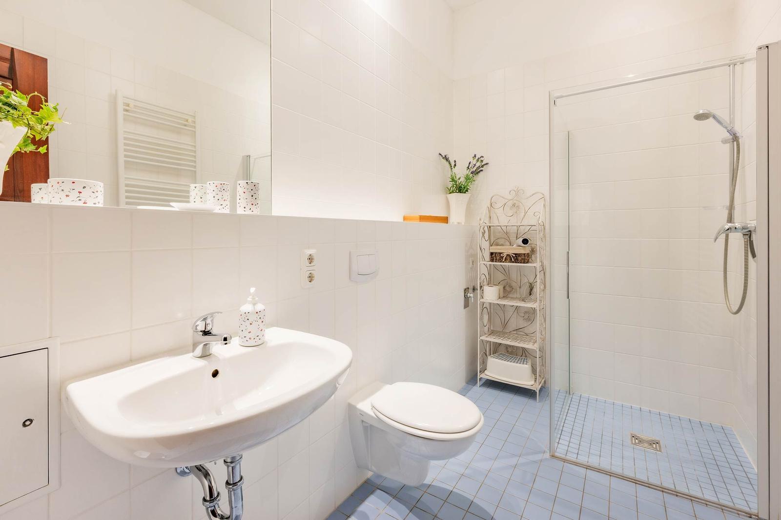 Badezimmer mit Dusche, Waschbecken und Toilette. Weiße Fliesen und blauer Bodenbelag.