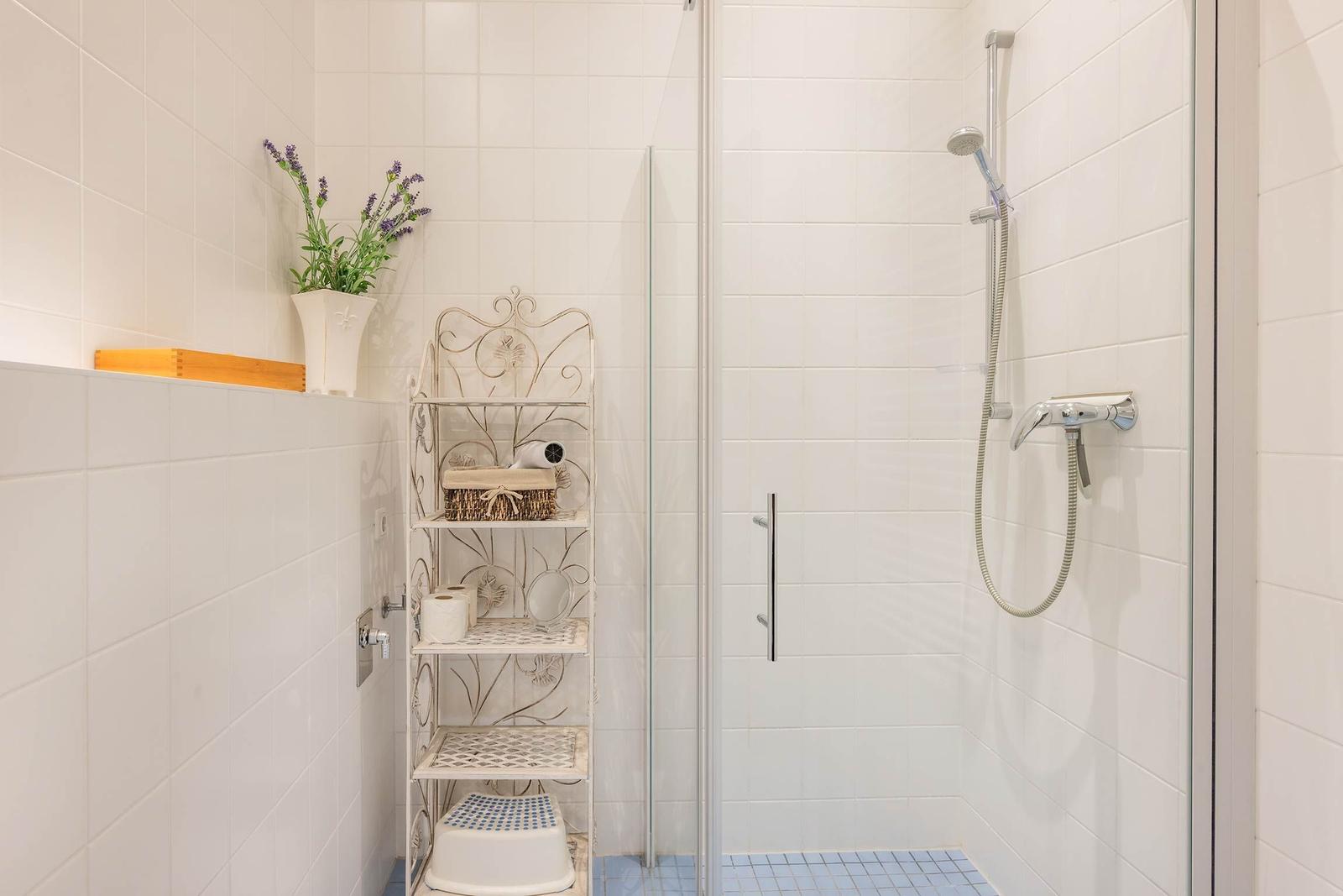Dusche mit Glaswand, Duschbrause und Regal mit Handtuch und Blumen.