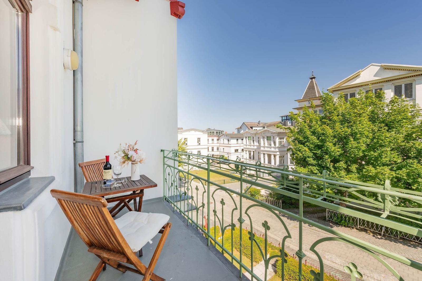 Balkon mit Tisch, Stühlen und Wein. Blick auf grüne Bäume und Gebäude.