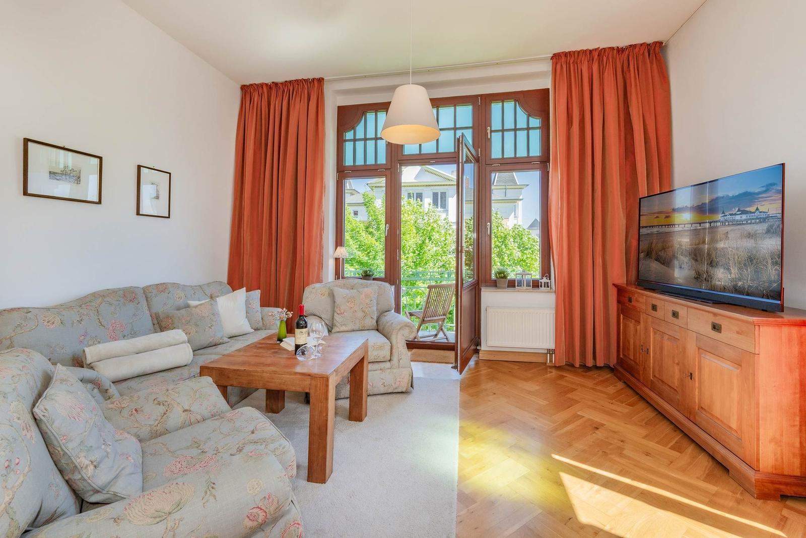 Wohnzimmer mit Sofa, Couchtisch und TV. Große Fenster mit Blick auf Balkon.
