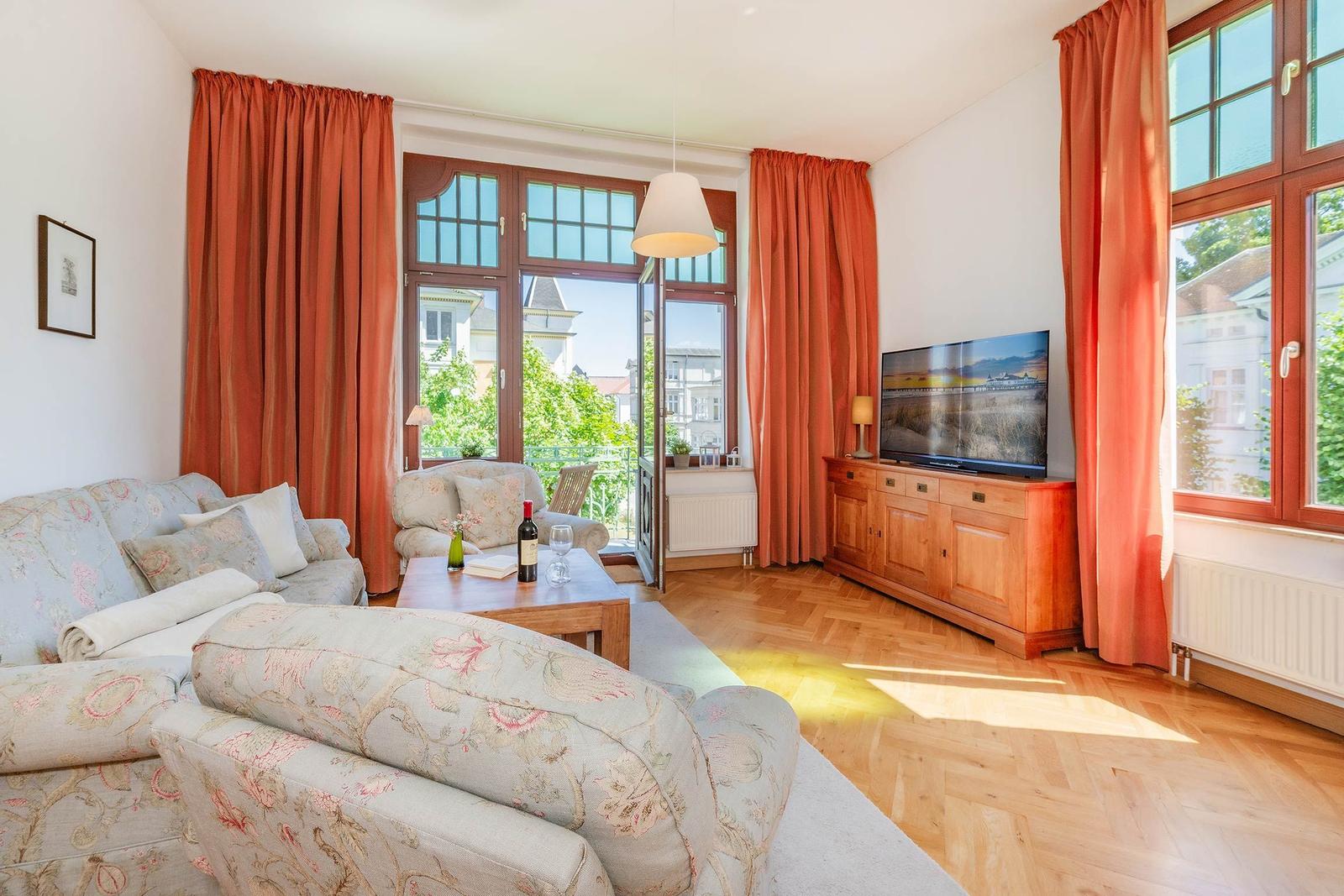 Wohnzimmer mit Sofa, Fernseher und Holzboden. Große Fenster mit orangefarbenen Vorhängen.