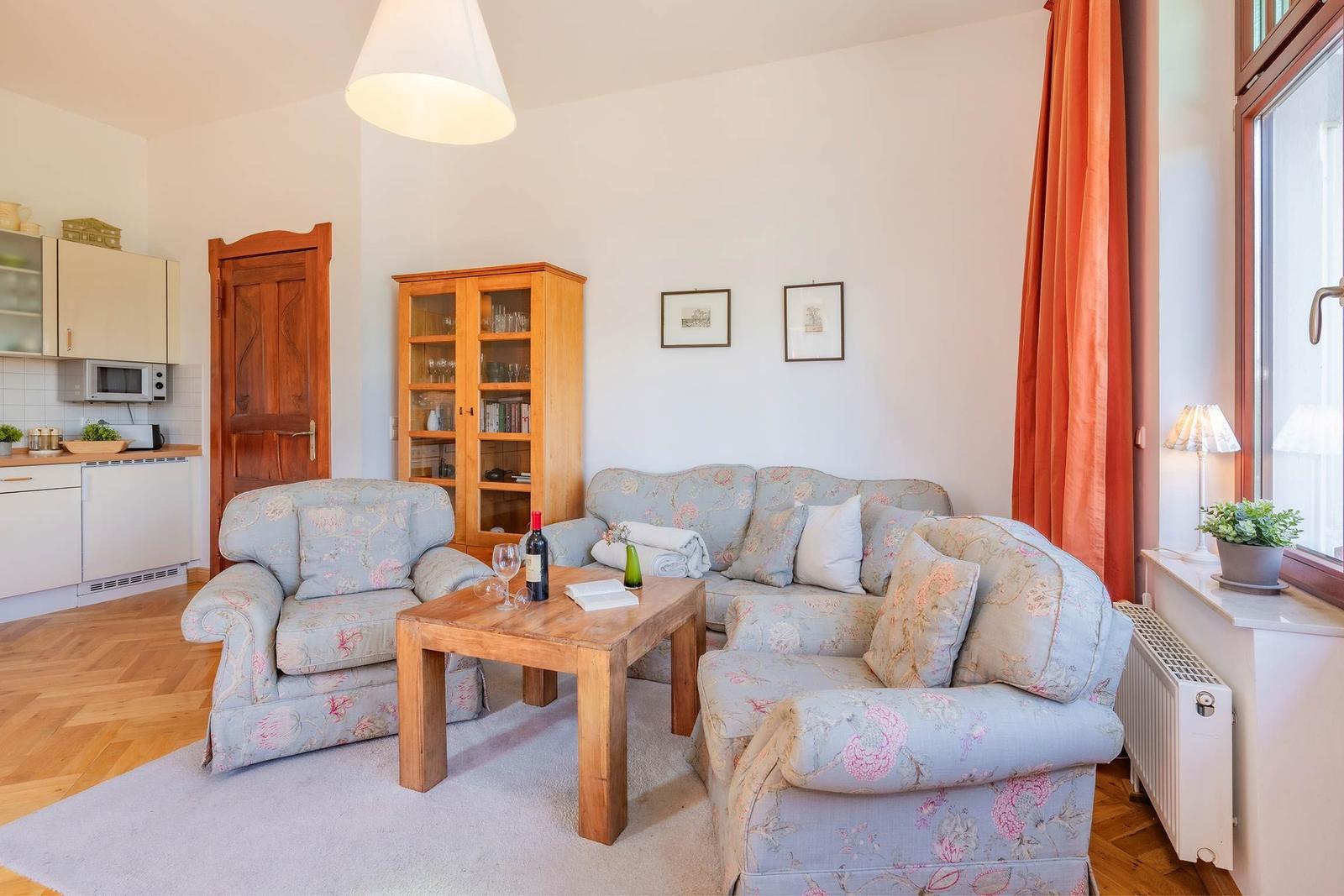 Wohnzimmer mit Sofa, Sessel und Holztisch. Fenster mit orangefarbenen Vorhängen.