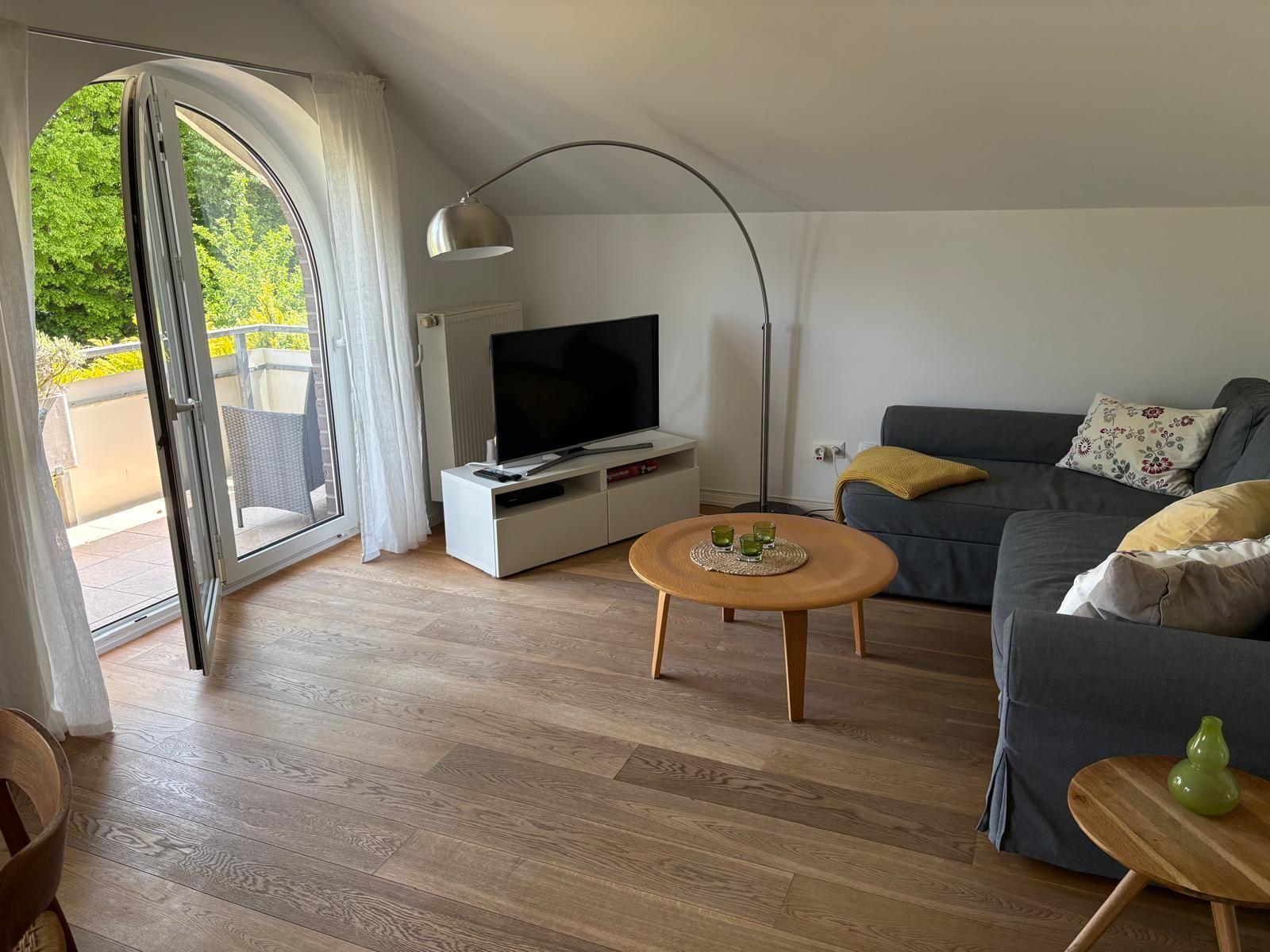 Wohnzimmer mit Sofa, Fernseher und Balkontür. Holzboden und runder Tisch.