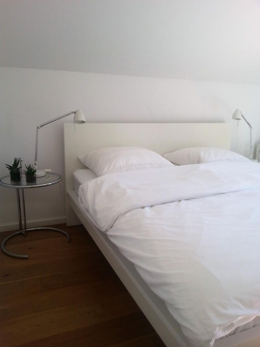 Ein Bett mit weißen Laken und Kissen. Links steht ein kleiner runder Tisch mit zwei Pflanzen und einer Lampe.