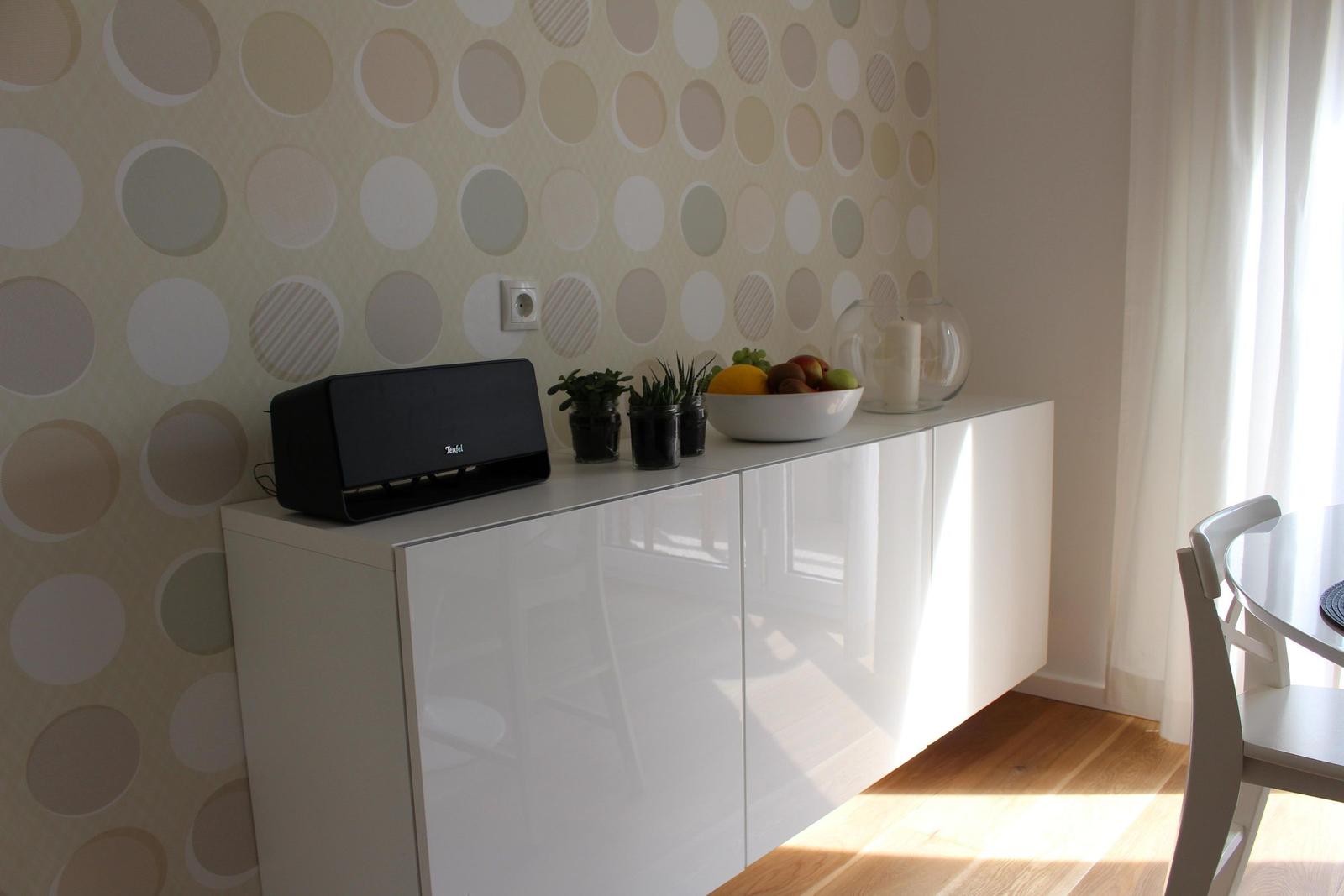 Weißer Sideboard mit Obstschale, Pflanzen und Lautsprecher vor gemustertem Wandbehang.