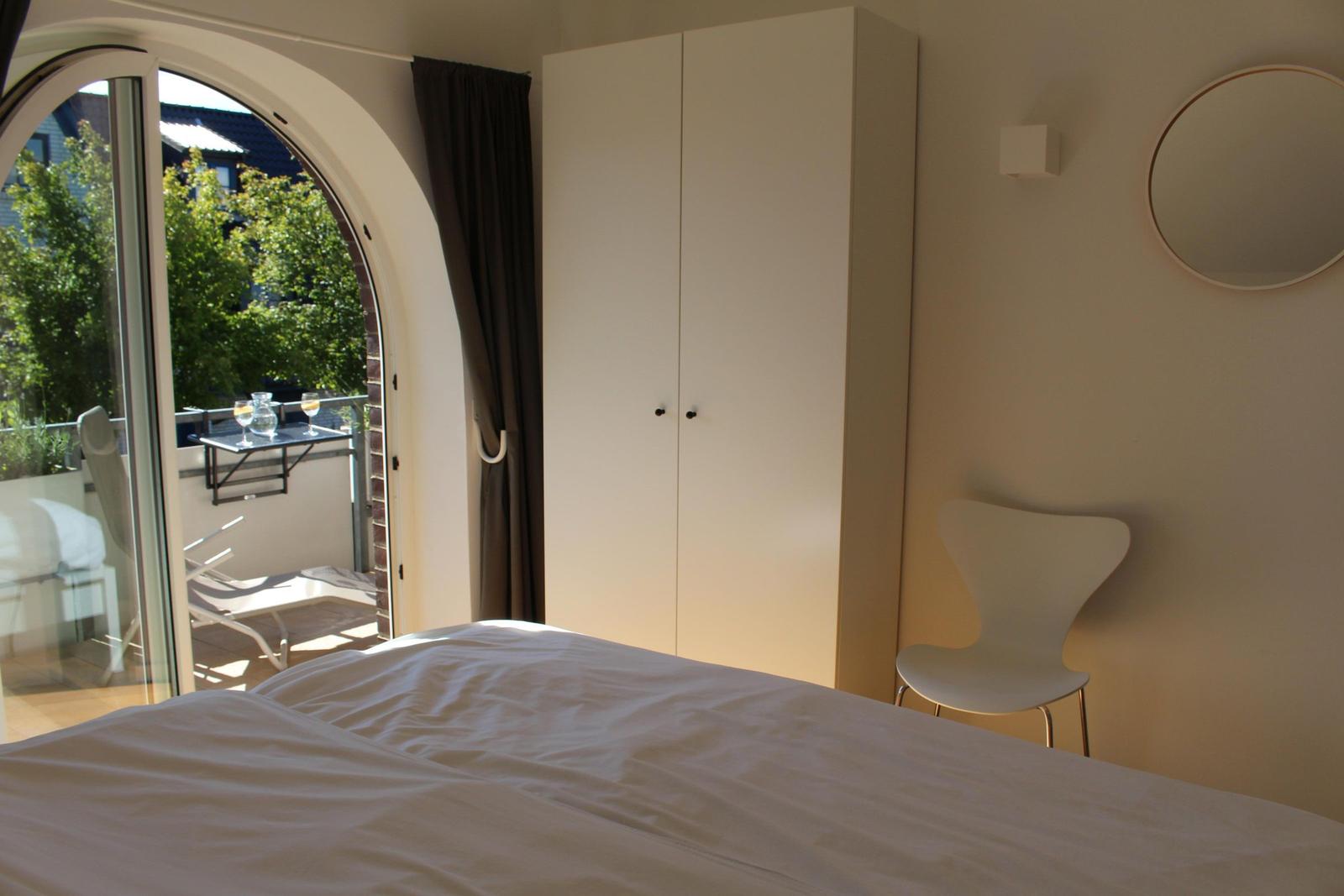 Schlafzimmer mit Bett, Kleiderschrank und Stuhl. Durch Fenster Zugang zu Balkon mit Tisch und Stühlen.