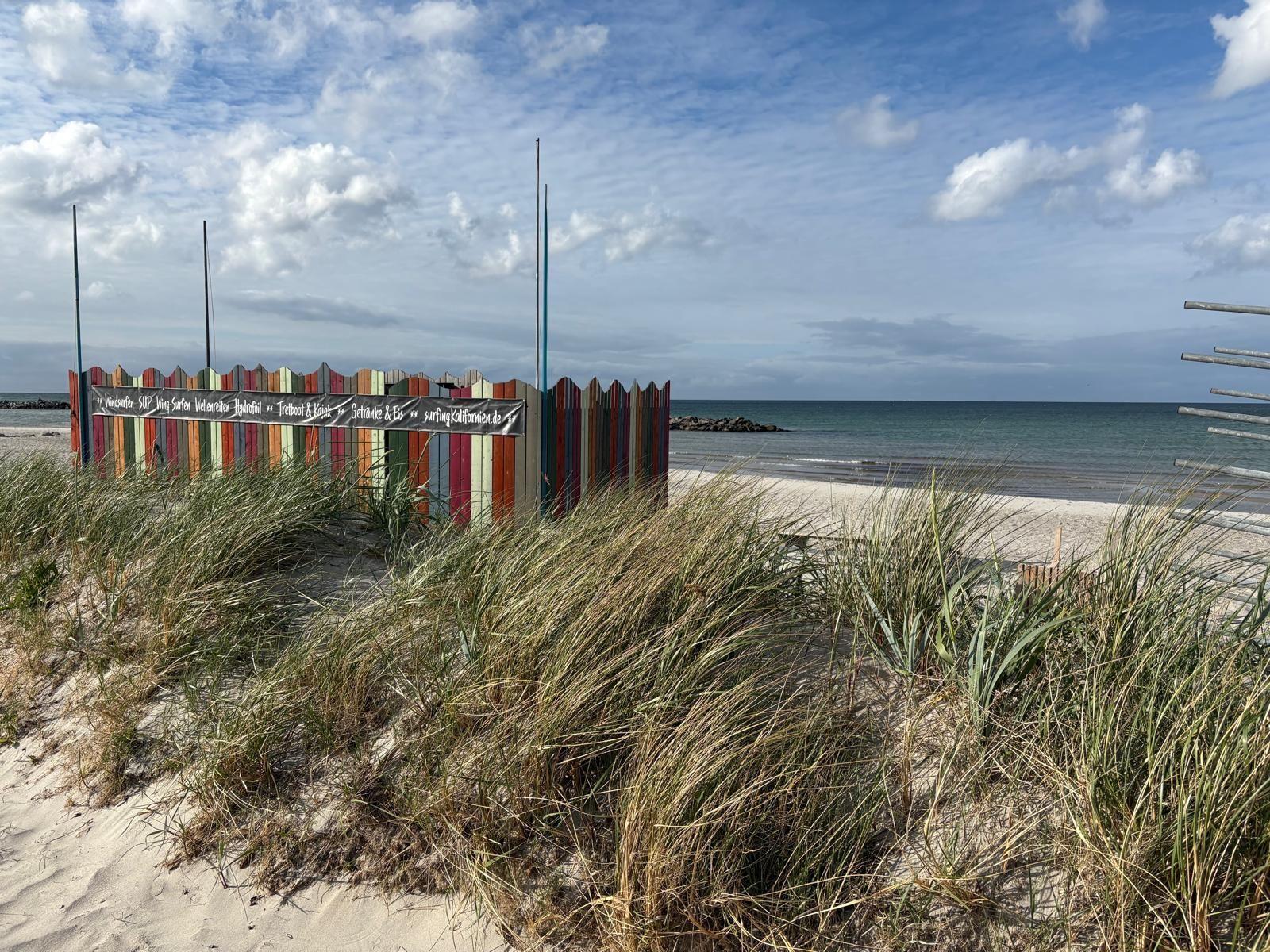 Farbiger Strandzaun mit Schild vor Sanddünen und Meer.
