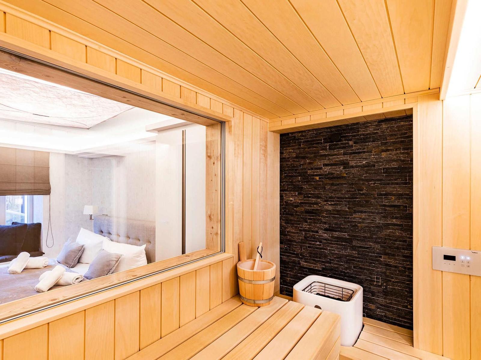 Sauna mit Holzverkleidung, Steinwand und Sitzbank. Durchgang zu Schlafzimmer mit Bett.