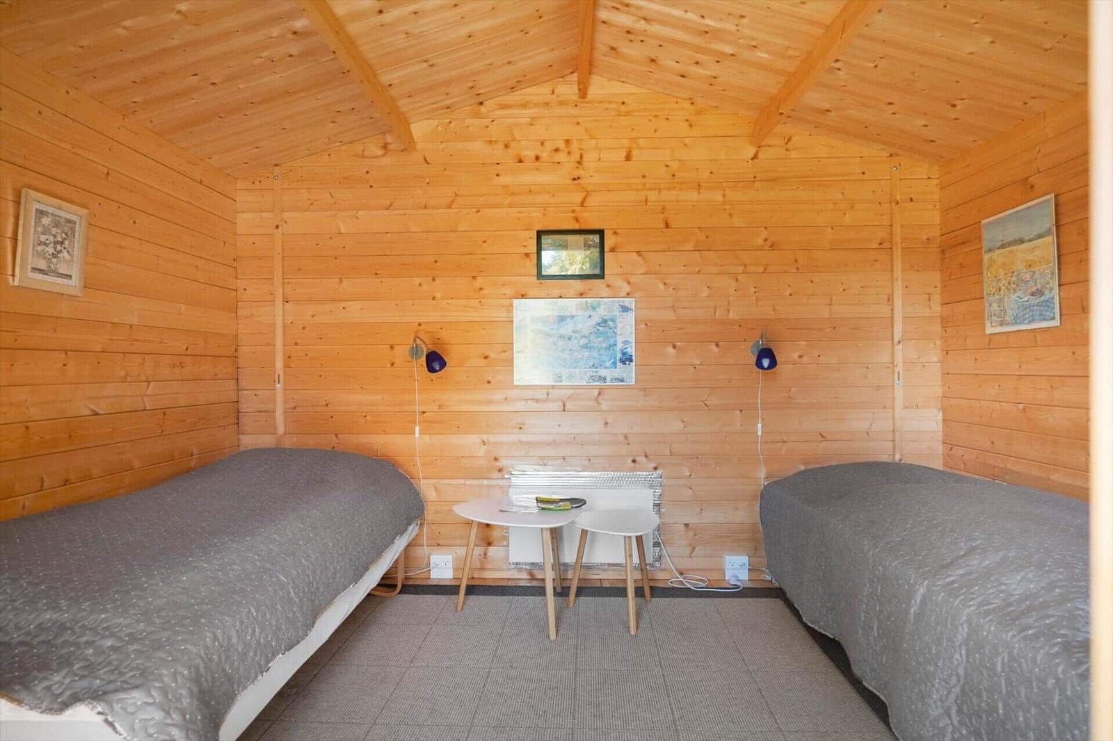 Zwei Betten mit grauer Bettwäsche, Tisch und Lampen in einem holzverkleideten Raum.