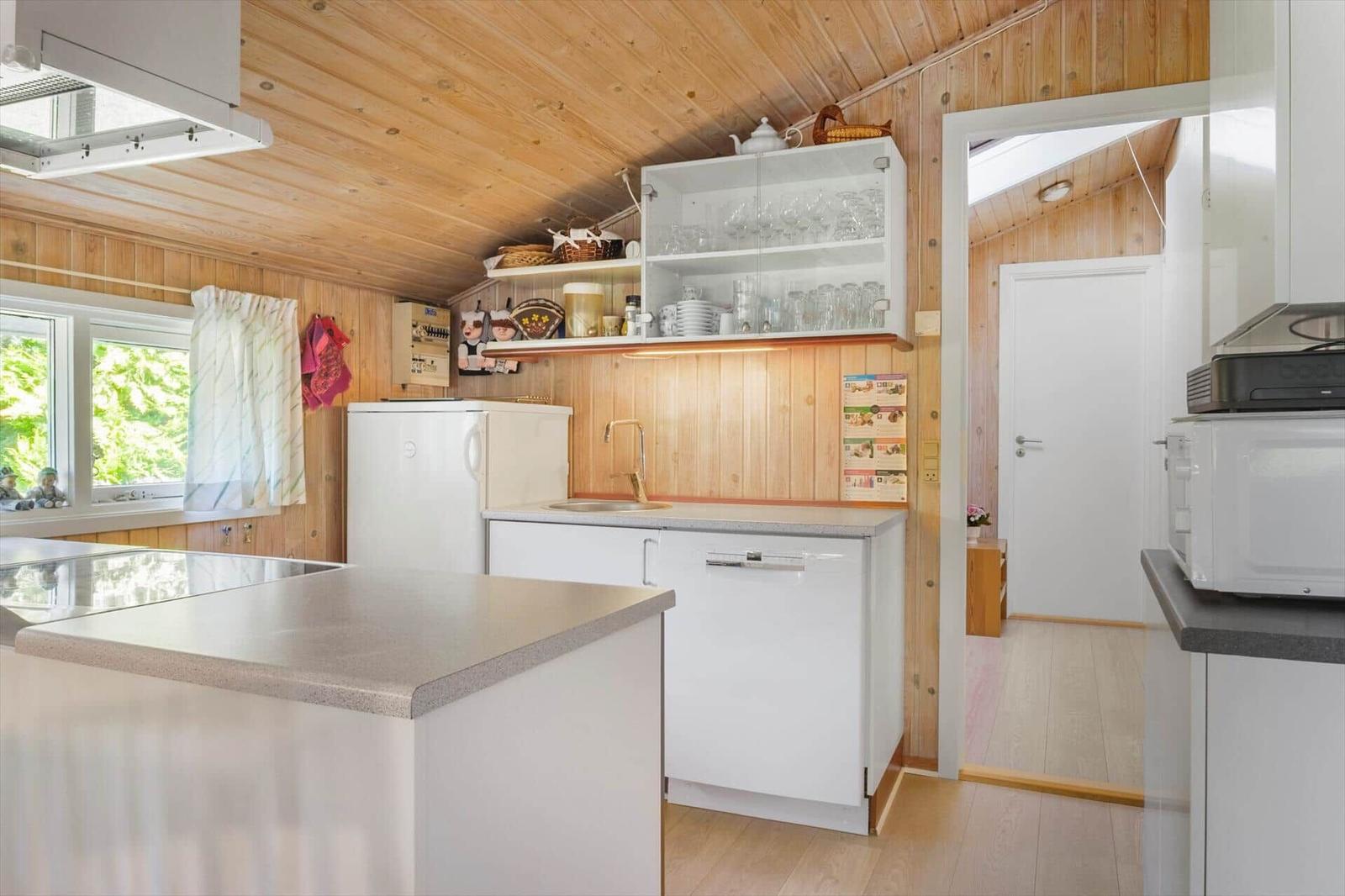 Küche mit Holzwand, weißen Schränken, Spüle und Kühlschrank. Durchgang zu einem anderen Raum.