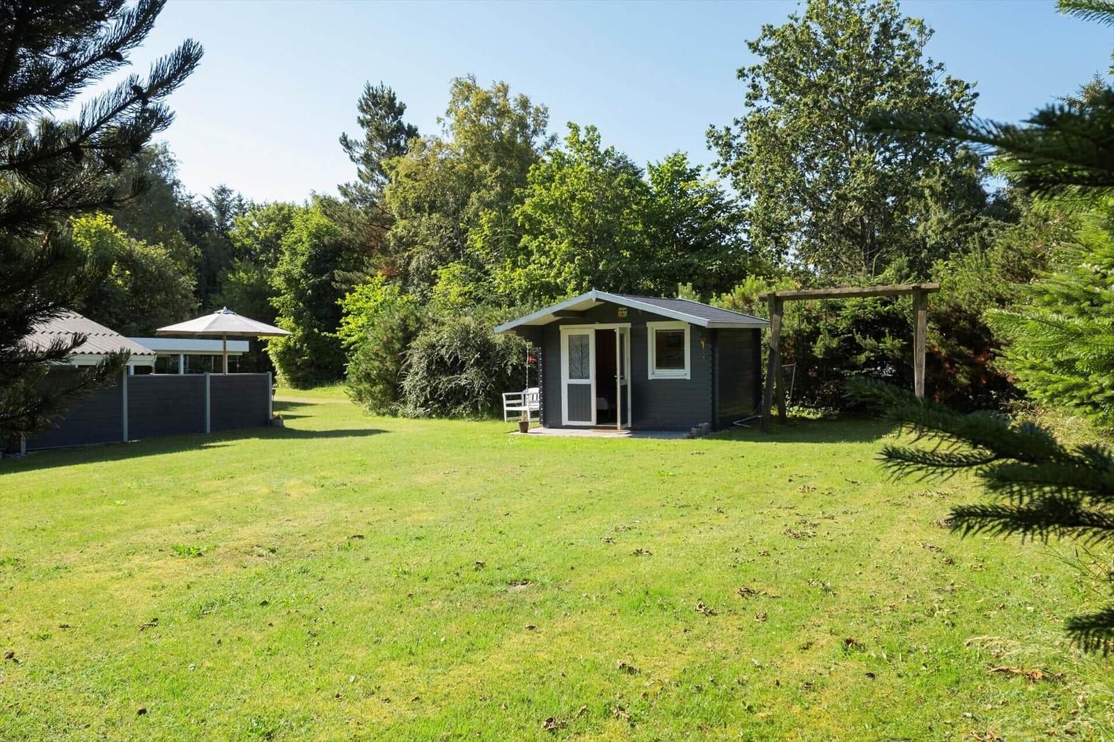 Ein kleines graues Haus mit weißer Tür und Fenster steht auf einer grünen Wiese umgeben von Bäumen.