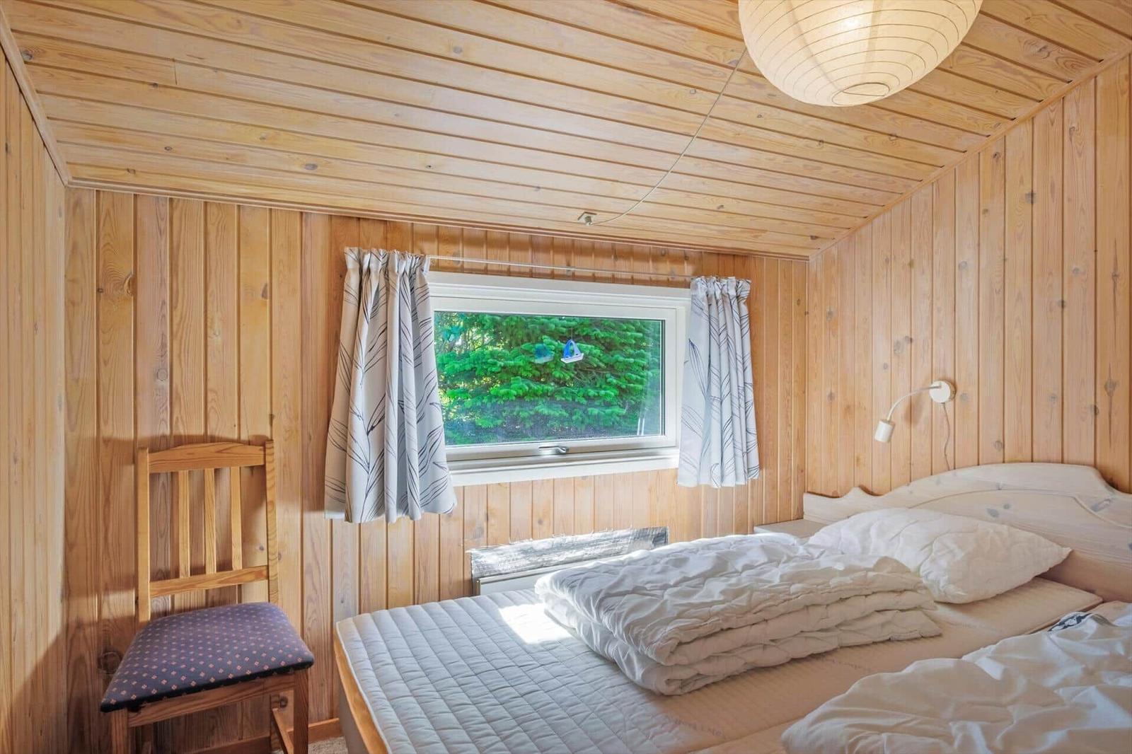 Schlafzimmer mit Holzwand, Bett, Stuhl und Fenster mit Blick auf Bäume.