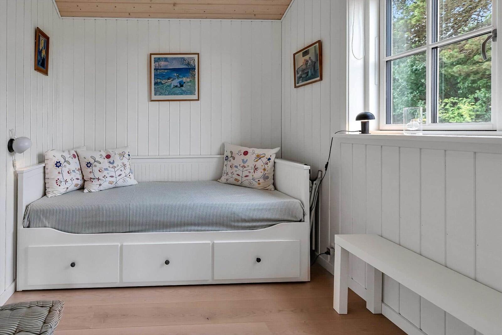 Ein weißes Bett mit drei Schubladen, Kissen und Wandbildern. Holzfußboden und Fenster.