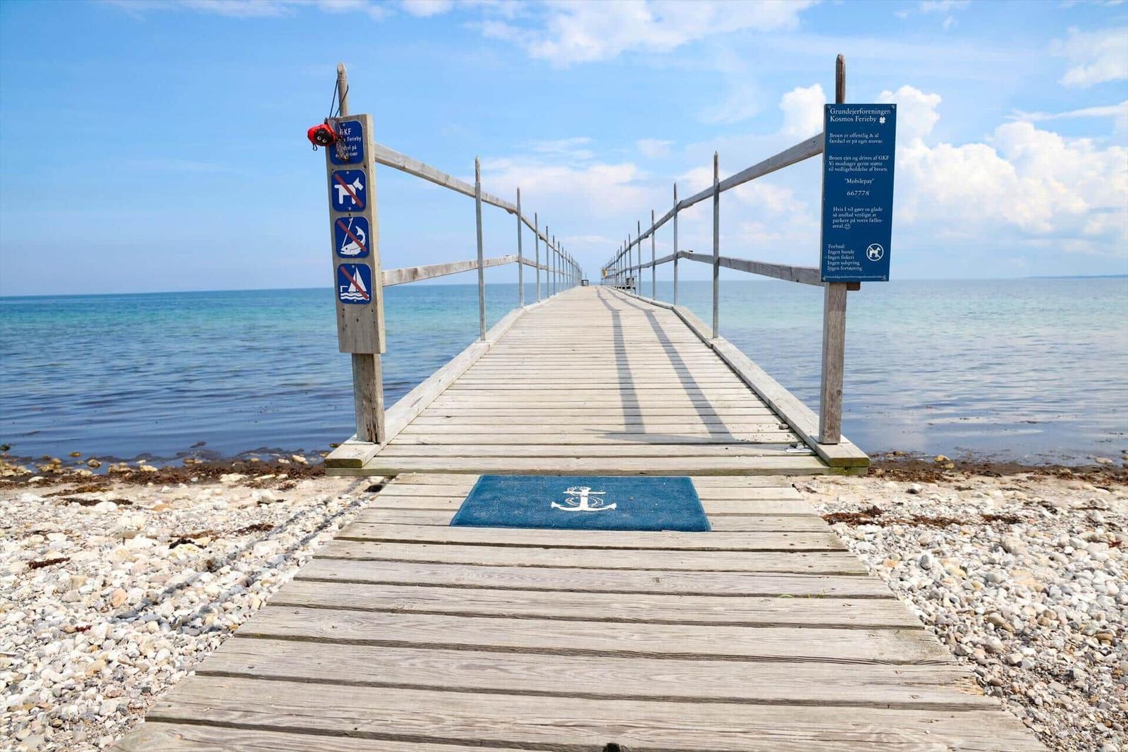 Holzsteg führt über Steinhügel zum Meer. Anker-Matte und Schilder sind sichtbar.