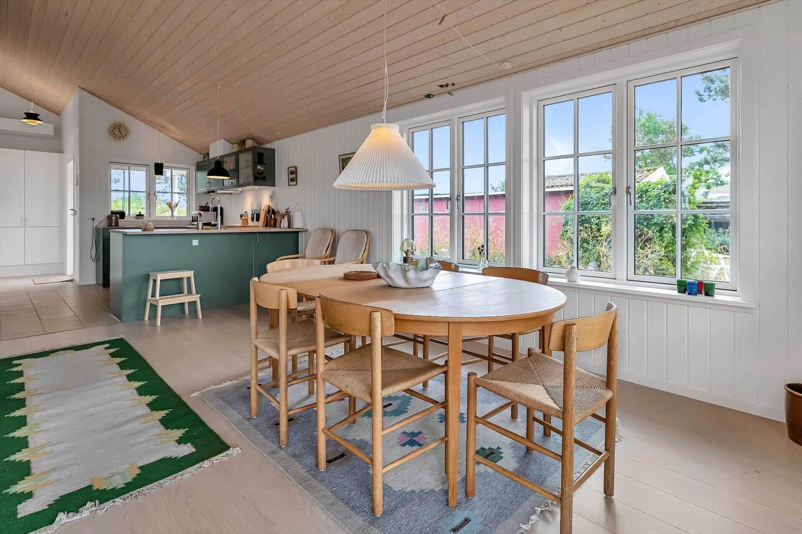 Essbereich mit Holztisch und Stühlen. Küche mit grünem Schrank. Große Fenster mit Blick auf Garten.