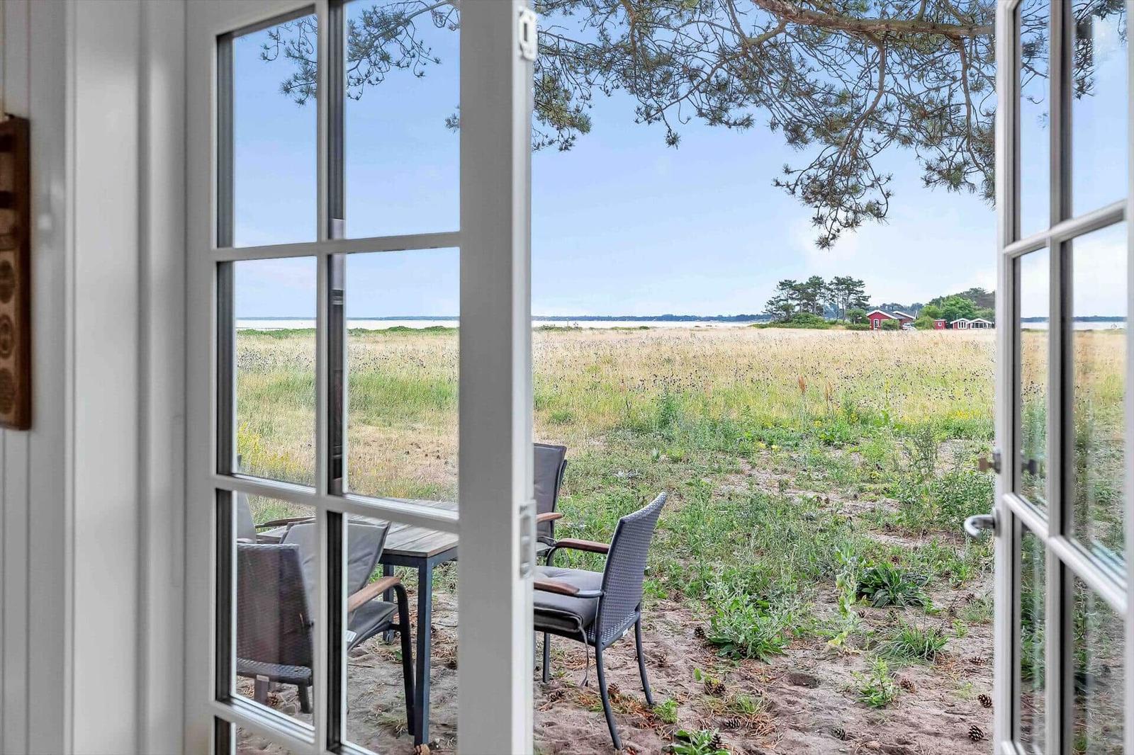 Durch offene Fenster blickt man auf einen sandigen Außenbereich mit Tisch und Stühlen.