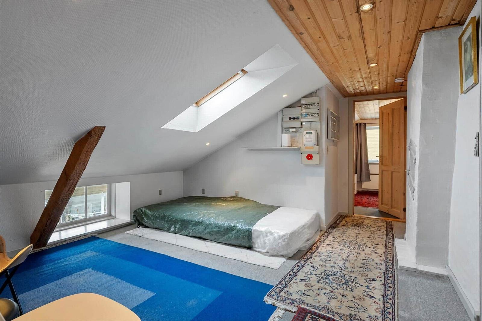 Dachgeschosszimmer mit Bett, Dachfenster und Holzdecke. Teppich und Fenster sichtbar.