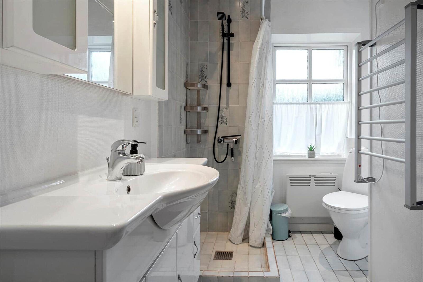 Badezimmer mit Dusche, Waschbecken und Toilette. Fenster mit Vorhang und Heizkörper.