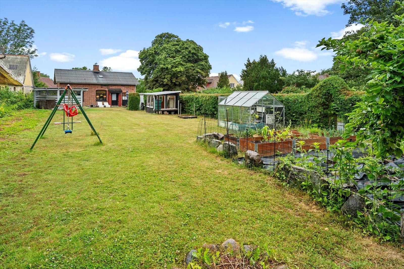 Grüner Garten mit Spielplatz, Gewächshaus und Beeten. Im Hintergrund steht ein Haus.