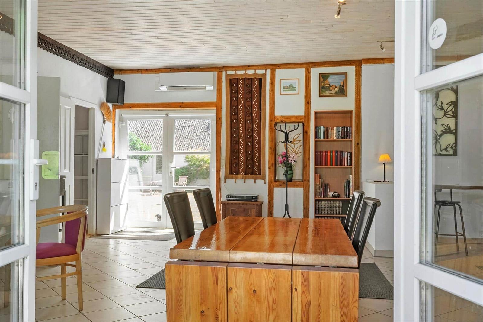 Dining room with wooden table, chairs, and bookcase. Passage to terrace with garden view.