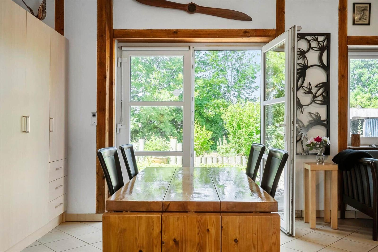 Dining room with wooden table, chairs, and view of green landscape through glass doors.