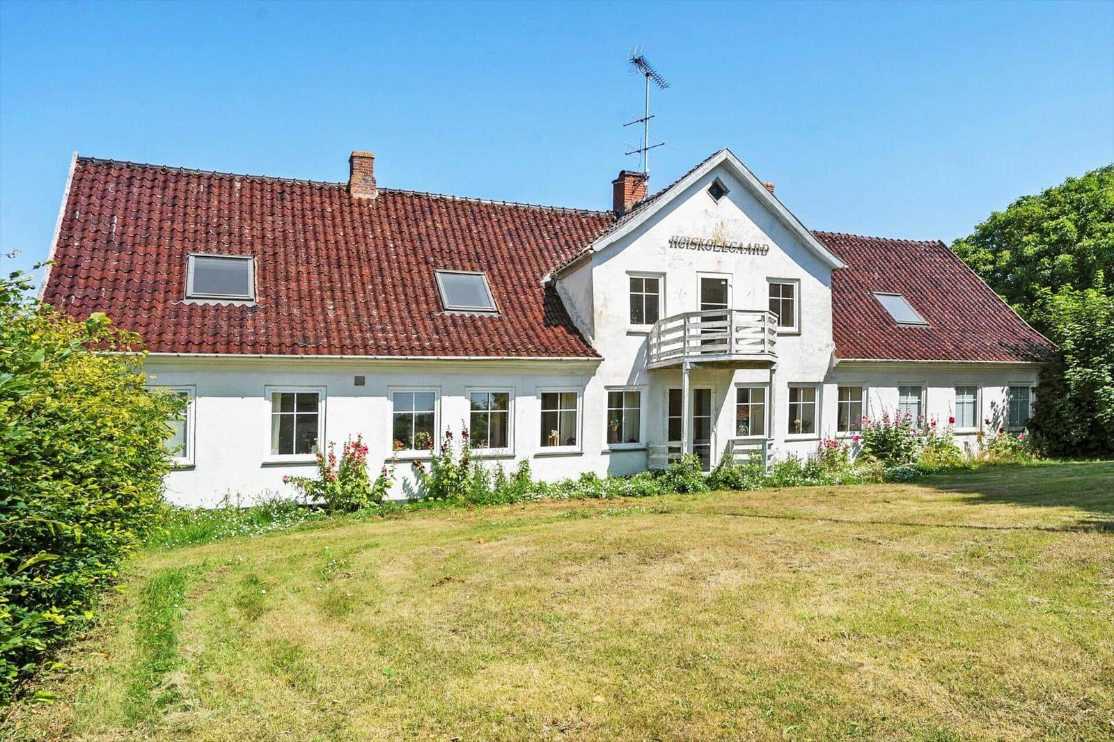 White holiday home with red roof and balcony. Surrounded by greenery with garden.