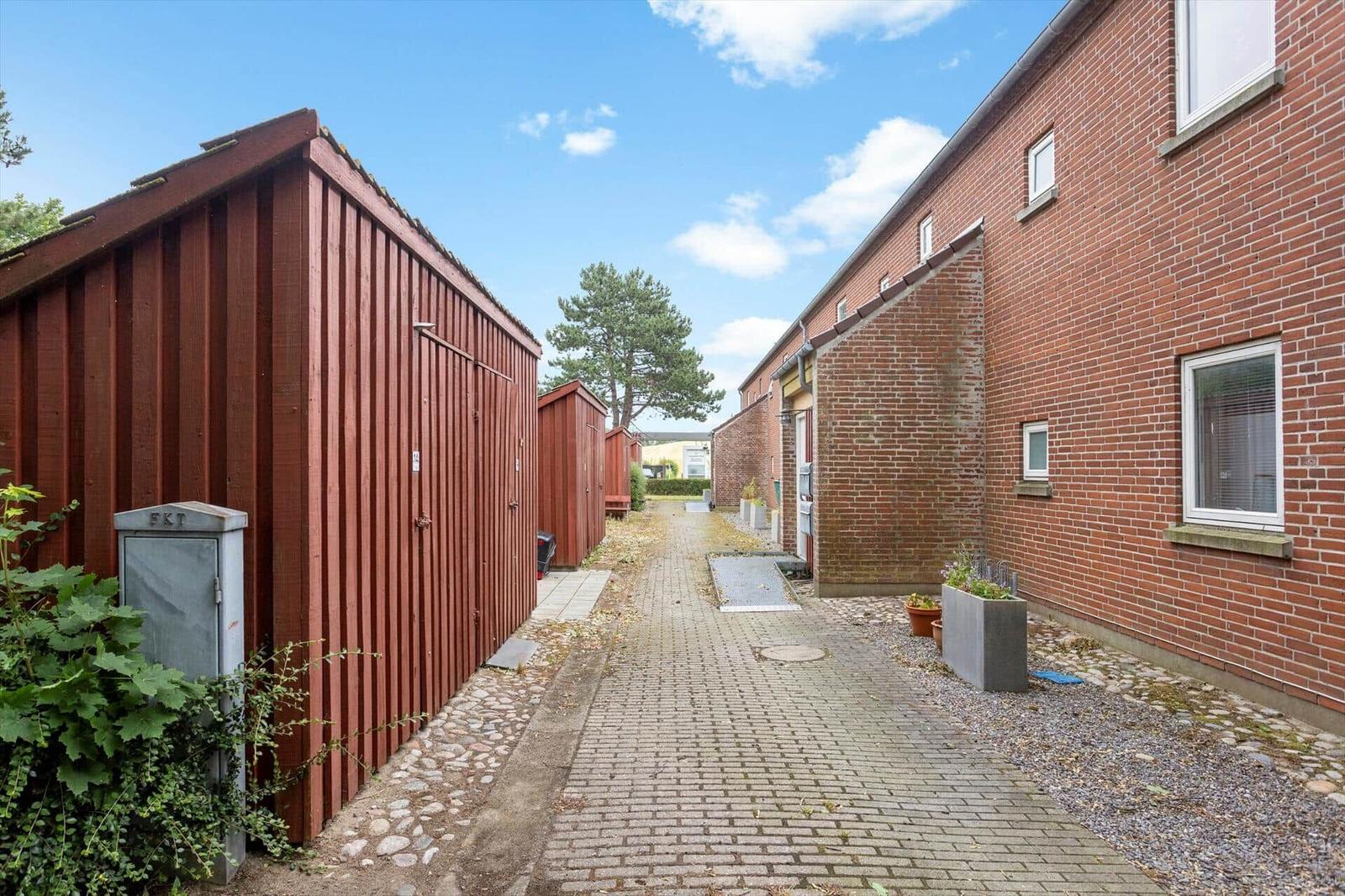Gehweg zwischen roten Holzcontainern und Backsteinhaus mit Fenstern.