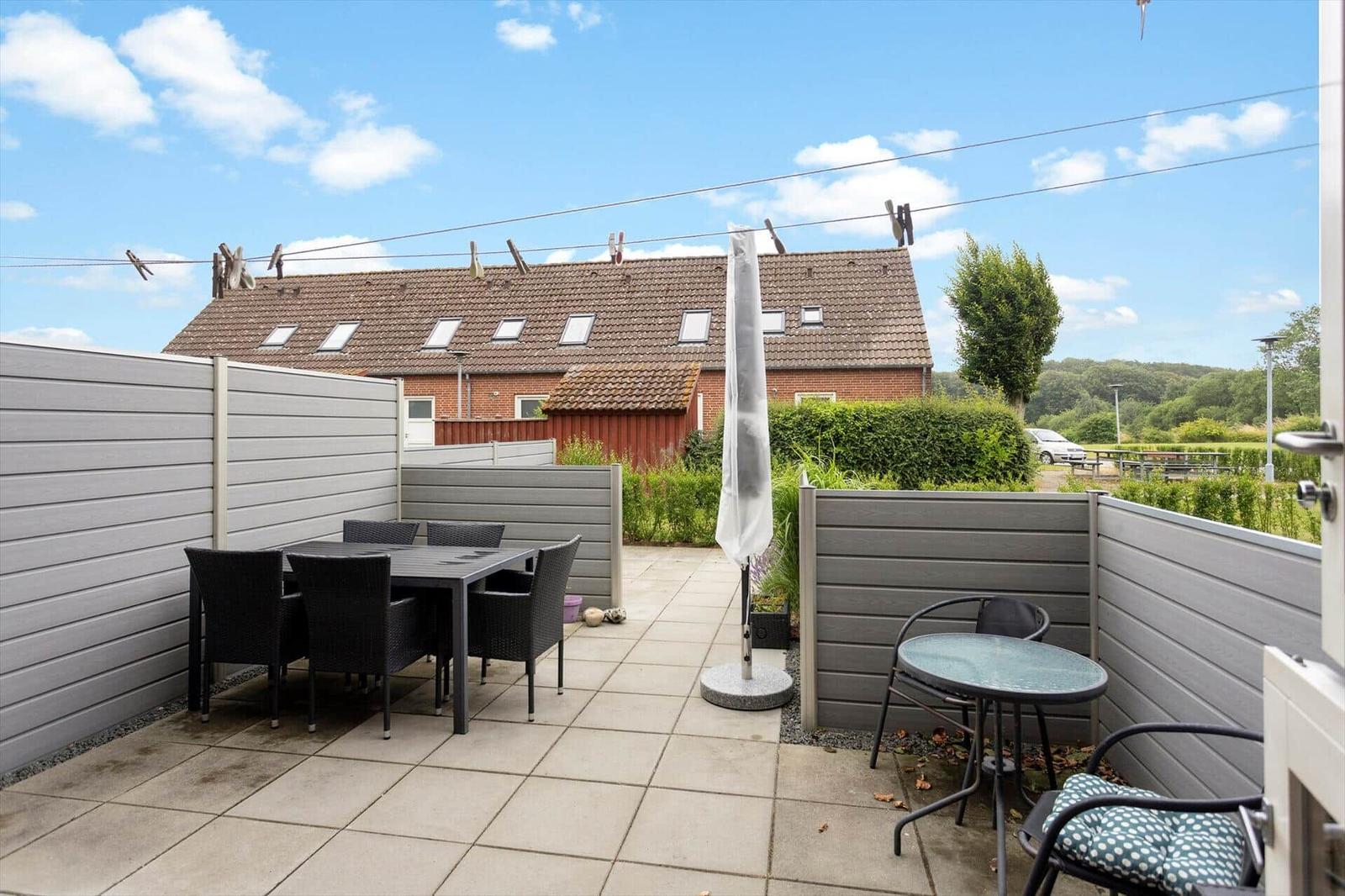 Terrasse mit Tisch, Stühlen und Sonnenschirm. Hinterhof mit Zaun und Blick auf Haus.