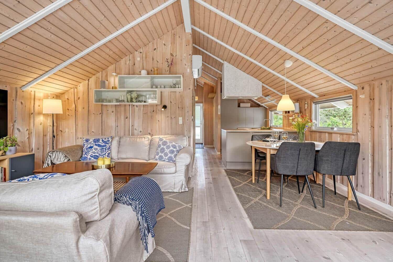 Wohn- und Essbereich mit Holzwand und Dachschrägen. Esstisch mit Stühlen und Küchenbereich.