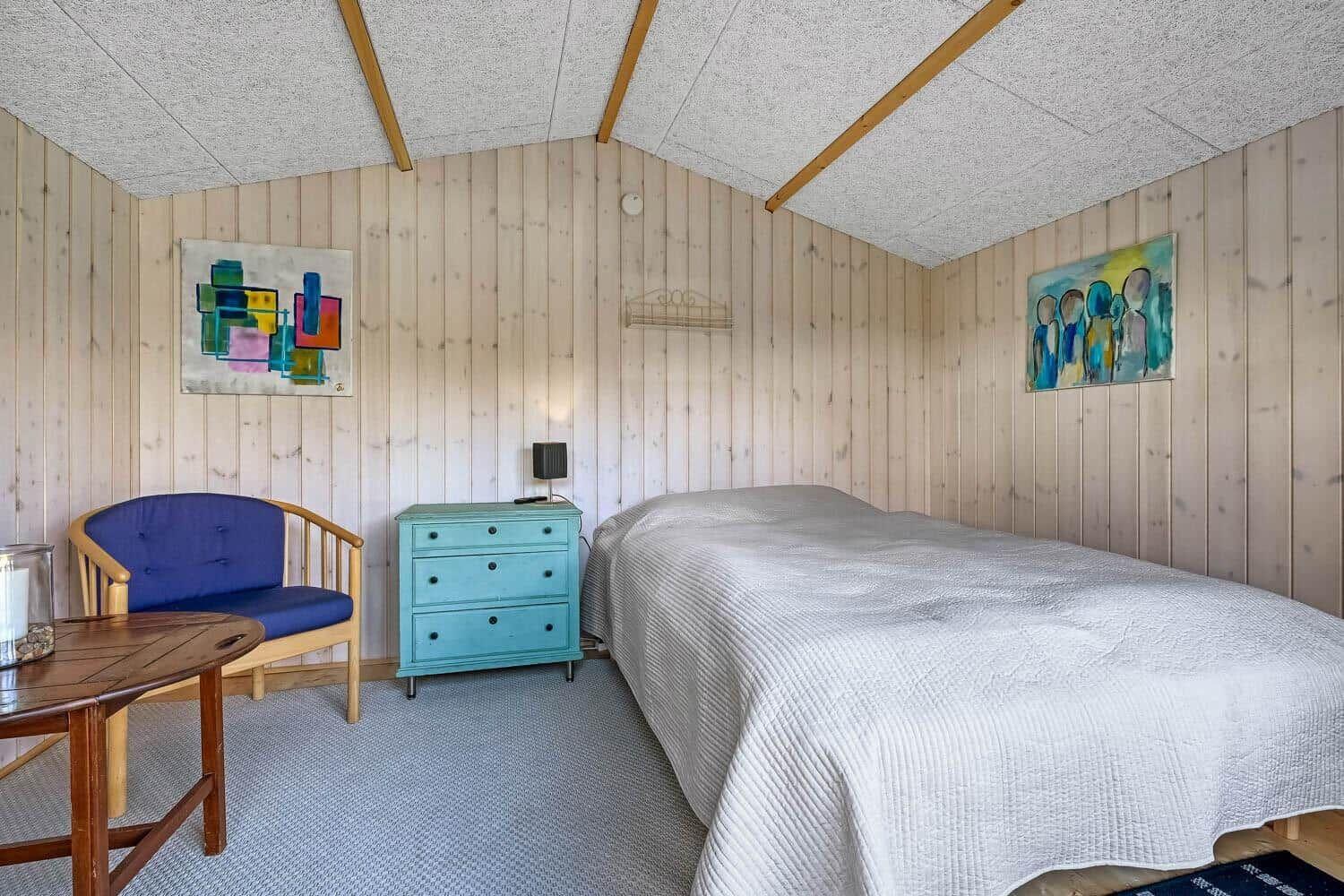 Schlafzimmer mit Bett, blauer Sitzgelegenheit, Schrank und Wandbildern.