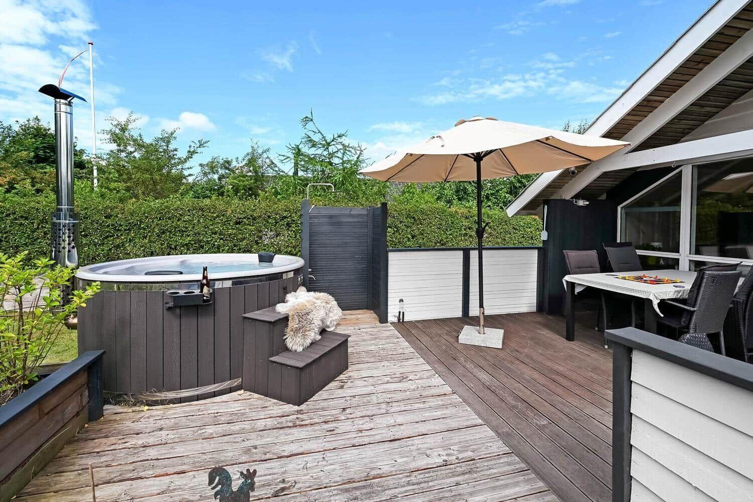 Terrasse mit Whirlpool, Sonnenschirm und Essbereich. Hintergrund: grüne Hecke und Haus.