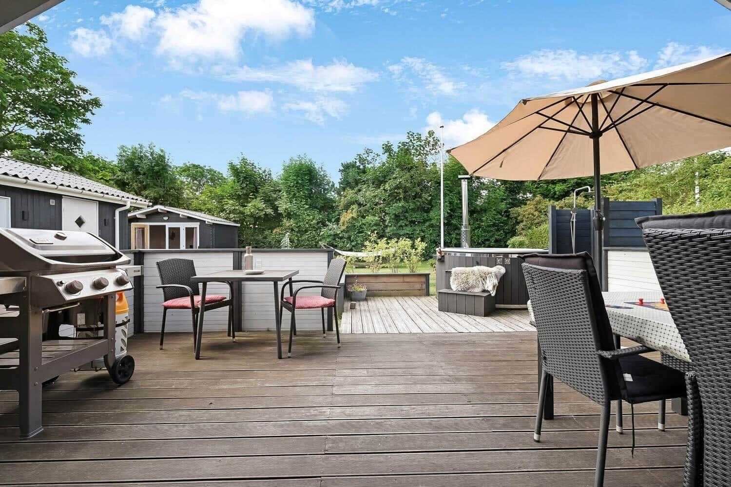 Terrasse mit Grill, Tisch, Stühlen und Sonnenschirm. Hintergrund: Bäume und Häuser.