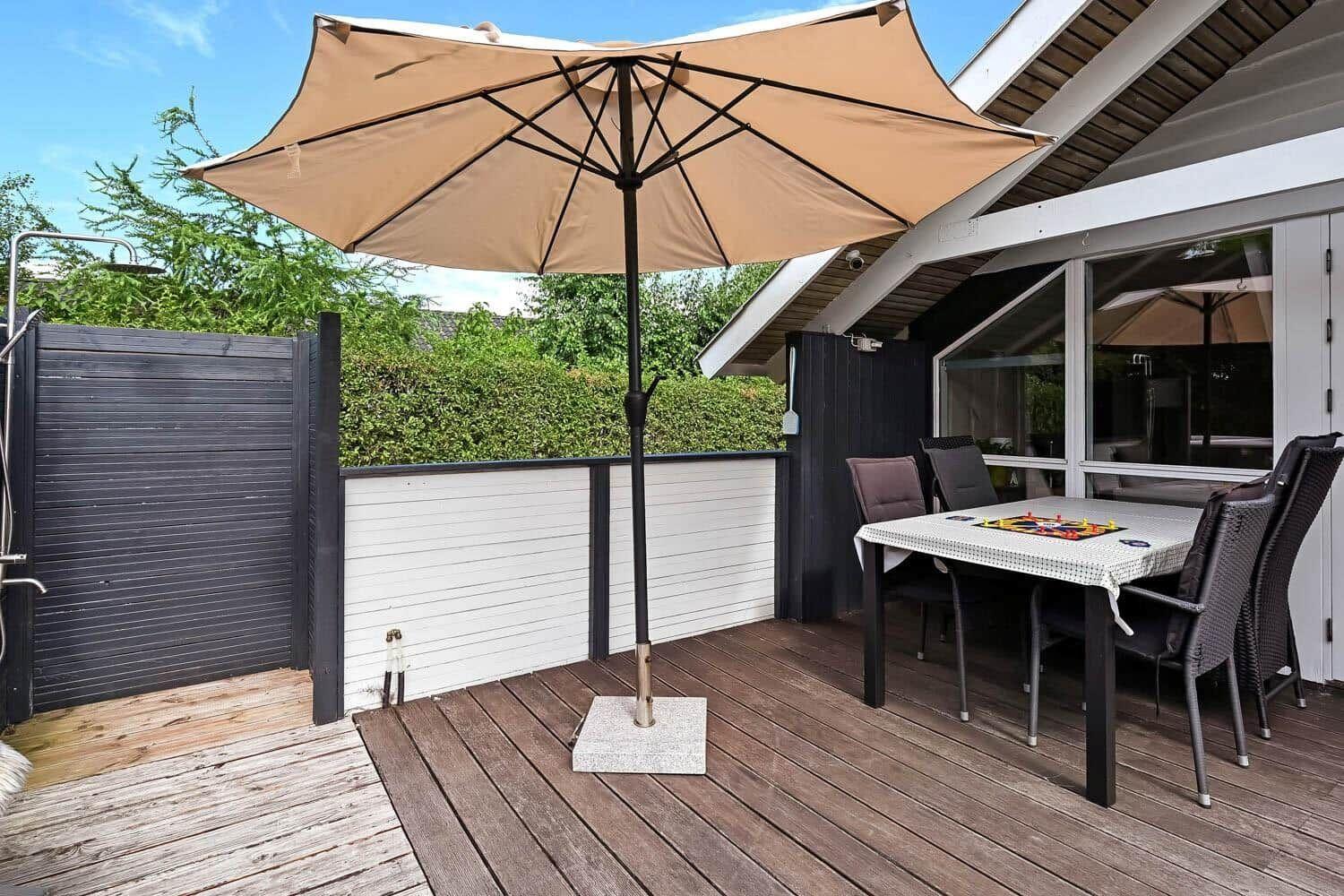 Terrasse mit Tisch, Stühlen und Sonnenschirm. Hintergrund: Holzgeländer und Grün.