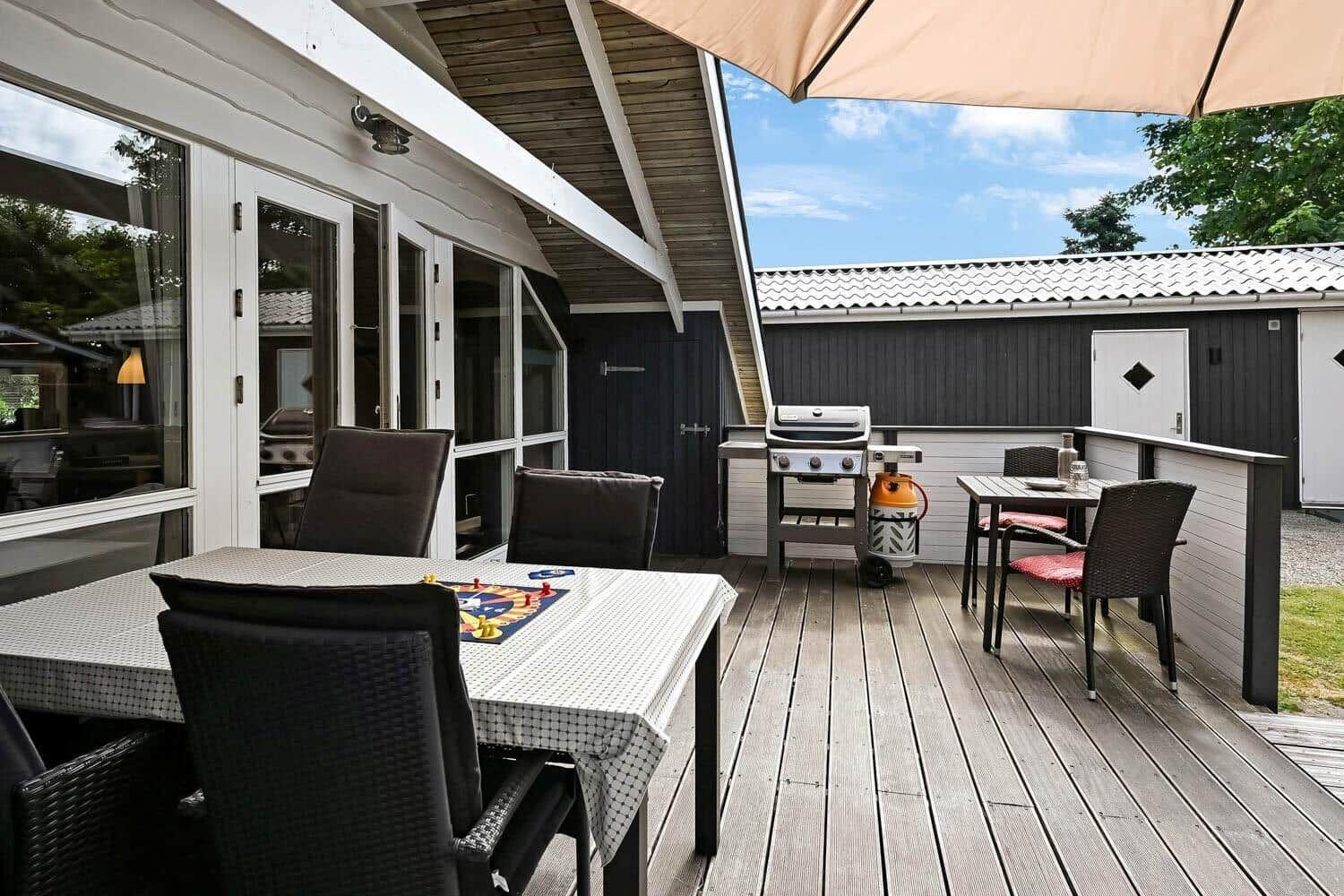 Terrasse mit Grill, Tisch und Stühlen unter Sonnenschutz. Holzdecke und Sicht auf Nebengebäude.