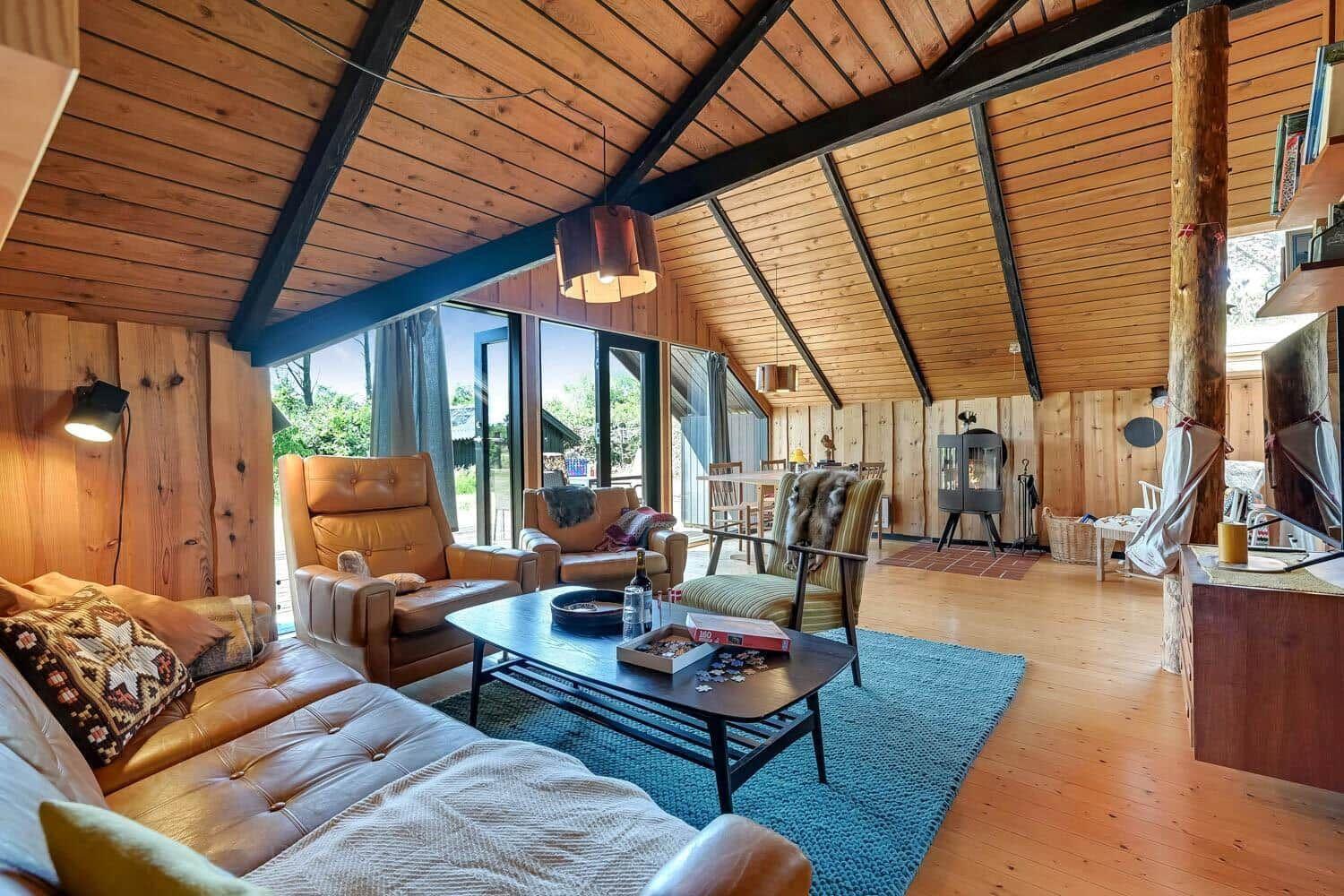 Wohnzimmer mit Holzwand, Sitzgelegenheiten und Kaminofen. Große Fensterfront mit Blick auf Garten.