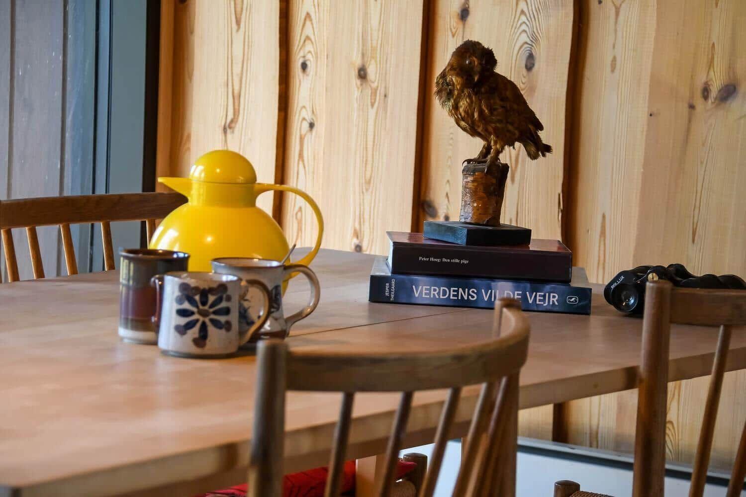 Holztisch mit Tassen, gelber Kanne und Buchstapel mit Vogelstatue.