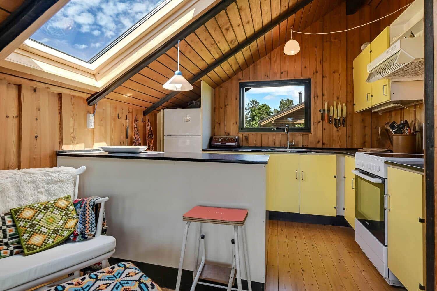 Küche mit gelben Schränken, Holzwand und Dachfenster. Sitzgelegenheit mit Kissen.