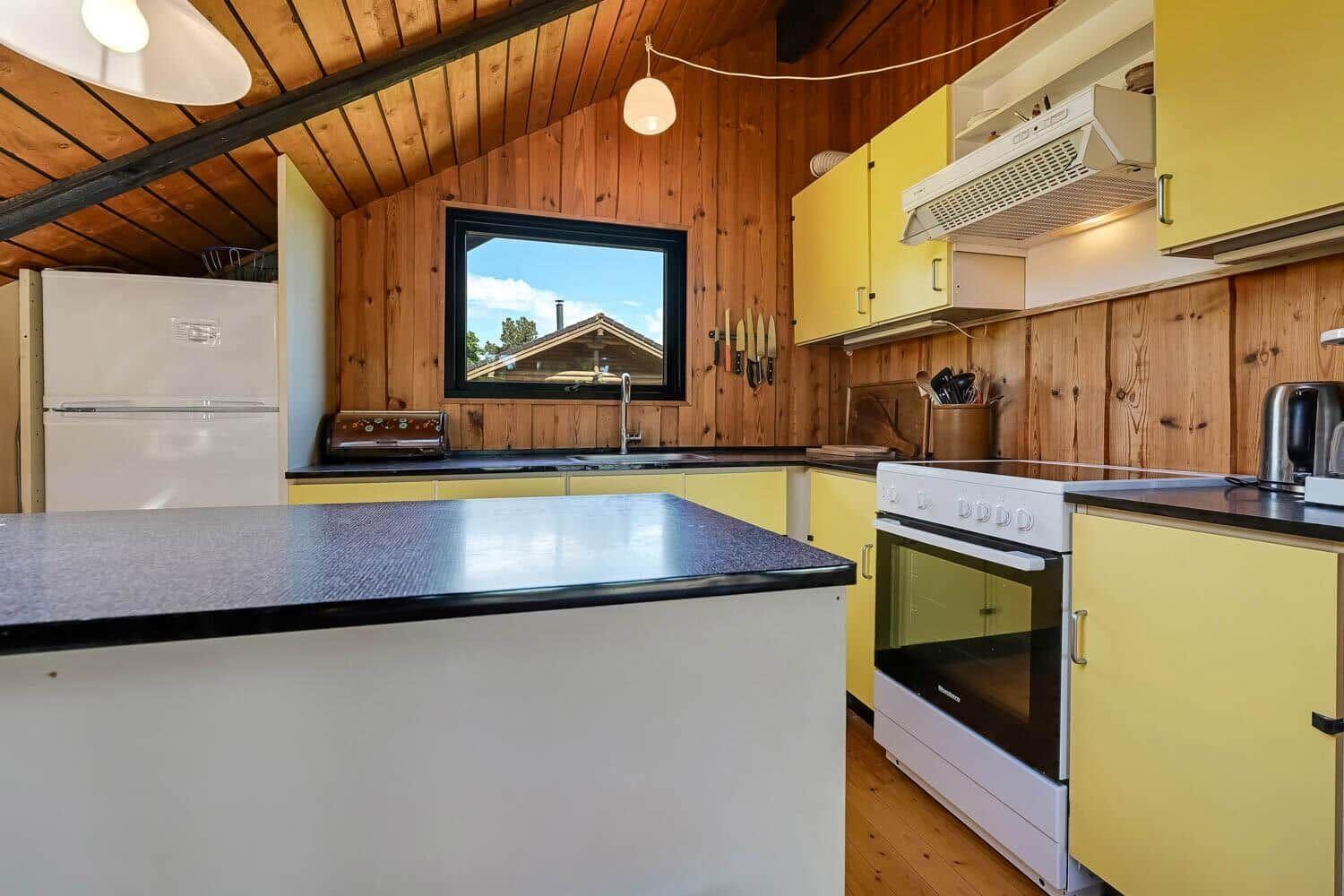 Küche mit gelben Schränken, weißen Geräten und Holzwand. Fenster zeigt Dach des Nachbarhauses.
