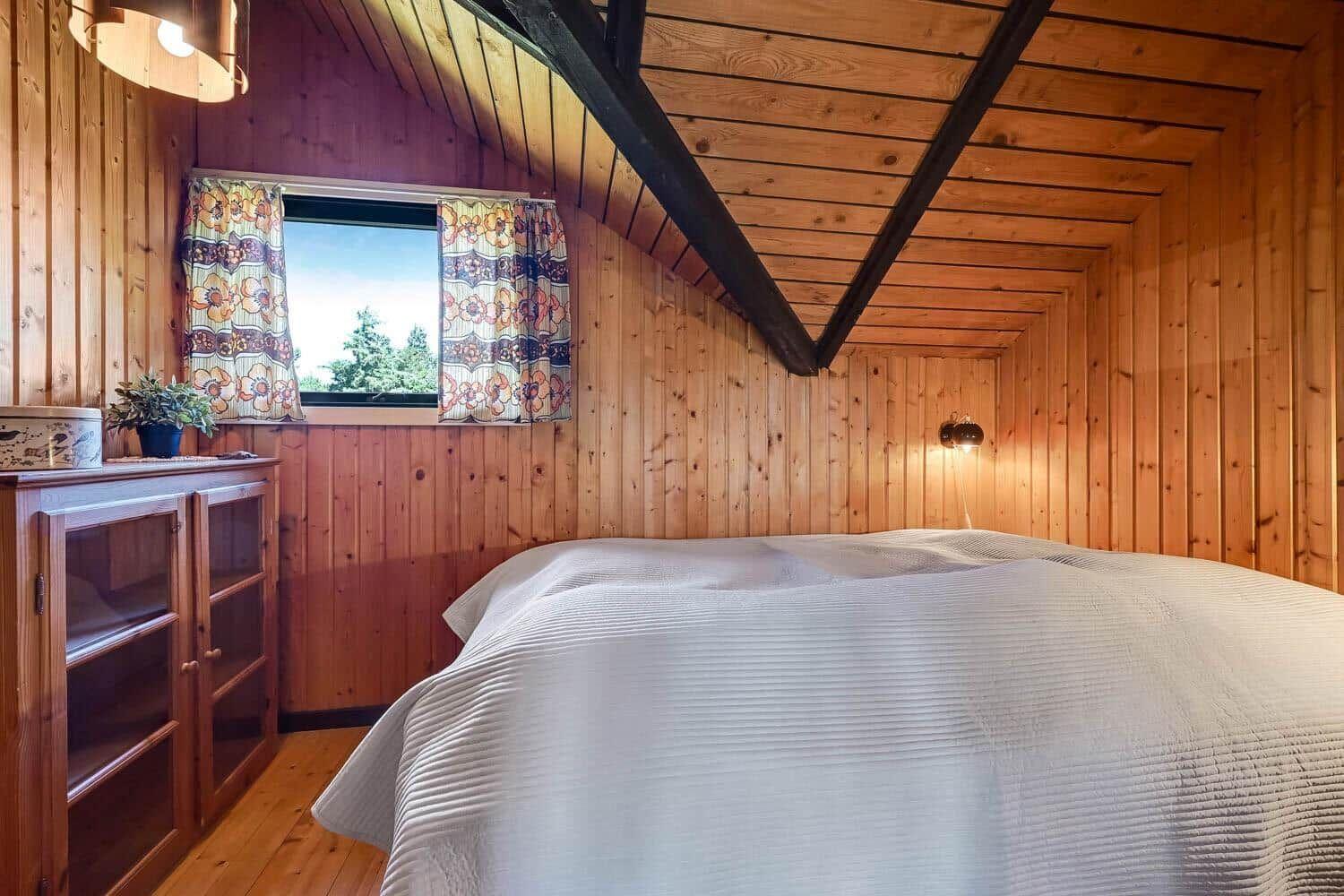 Schlafzimmer mit Holzwand, Bett und Fenster mit Vorhängen.