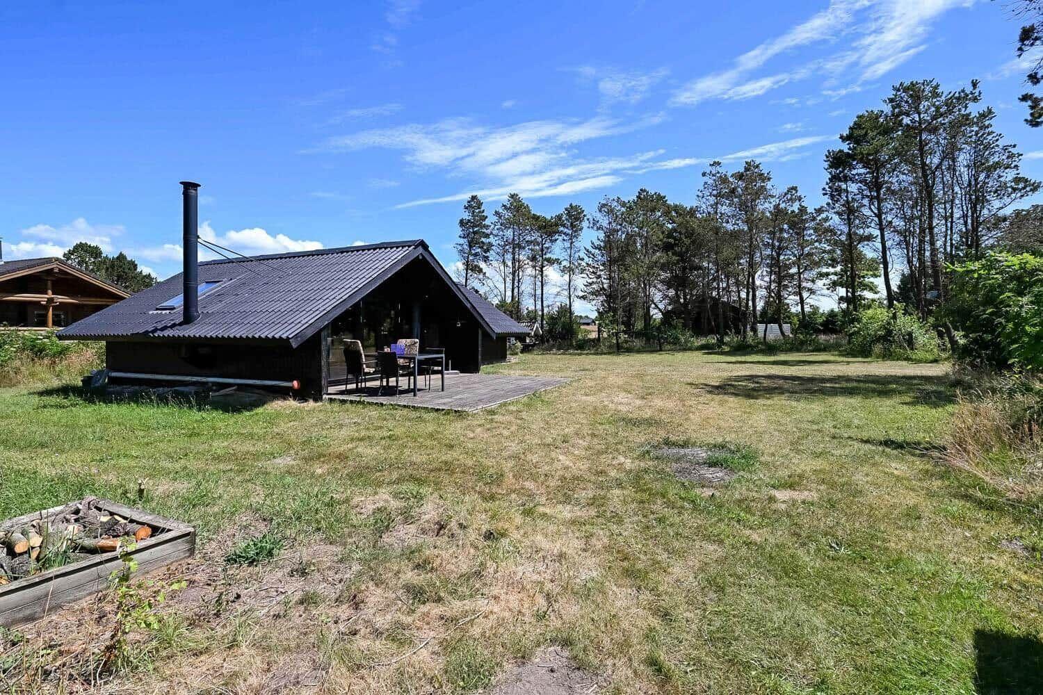 Hütte mit Terrasse, Grillplatz und Wald im Hintergrund unter blauem Himmel.