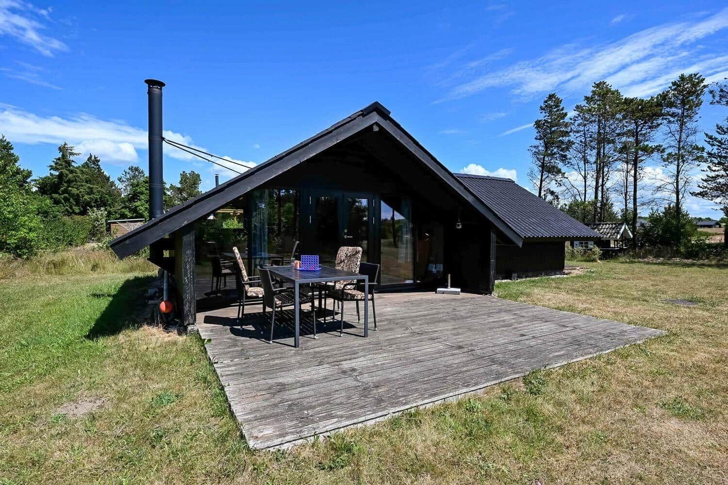 Moderne Holzhütte mit Terrasse und Grillplatz. Umgeben von Gras und Bäumen unter blauem Himmel.