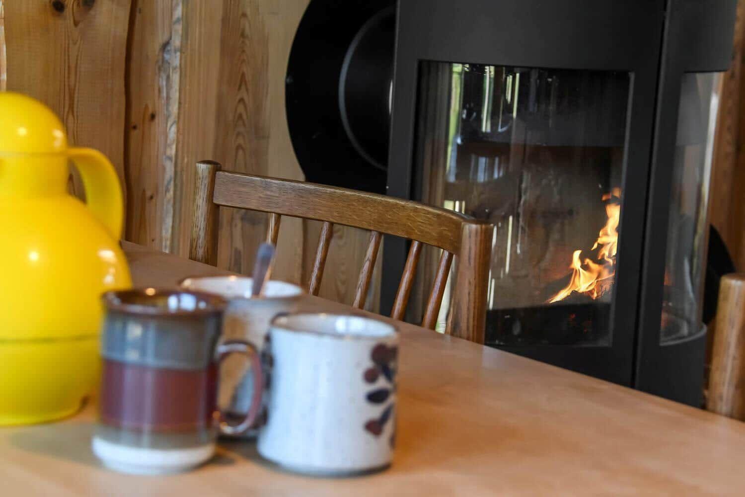Holztisch mit Tassen und Kanne vor Kamin mit brennendem Feuer.