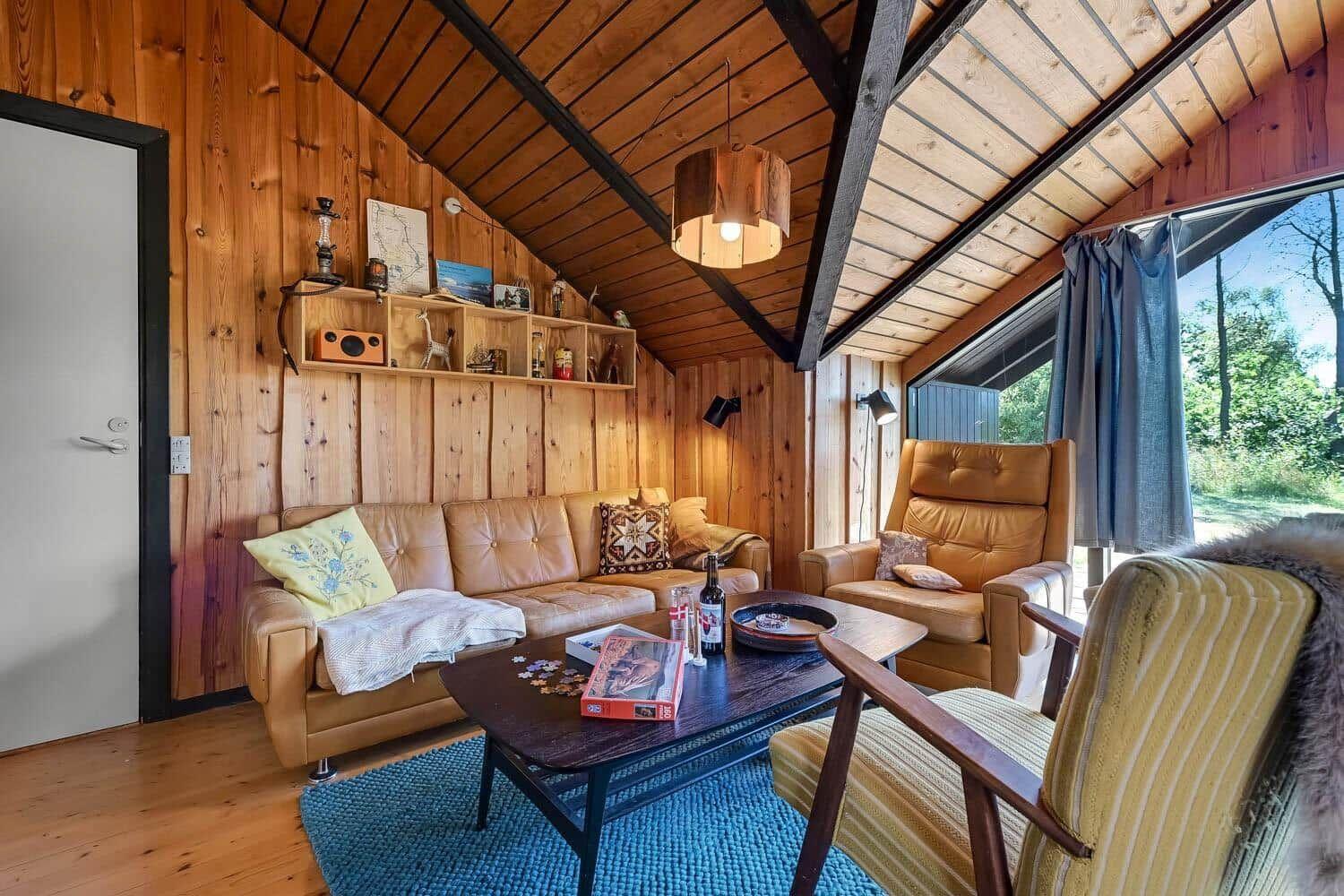 Wohnzimmer mit Holzwand, Sofa, Sessel und Tisch. Fenster mit Blick auf Wald.