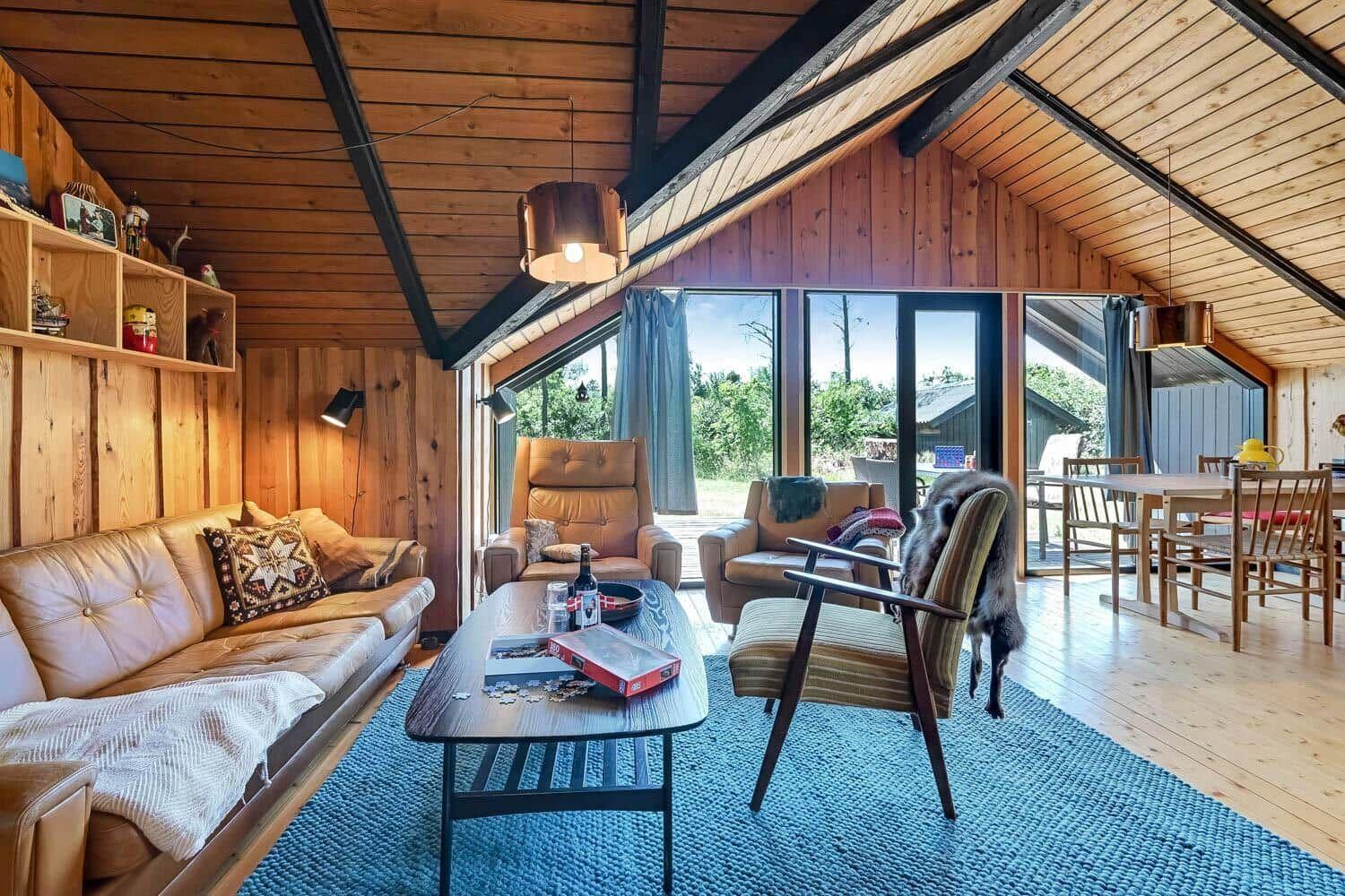 Wohn- und Essbereich mit Holzwand, Sofa, Stühlen und großem Fenster zur Außenseite.